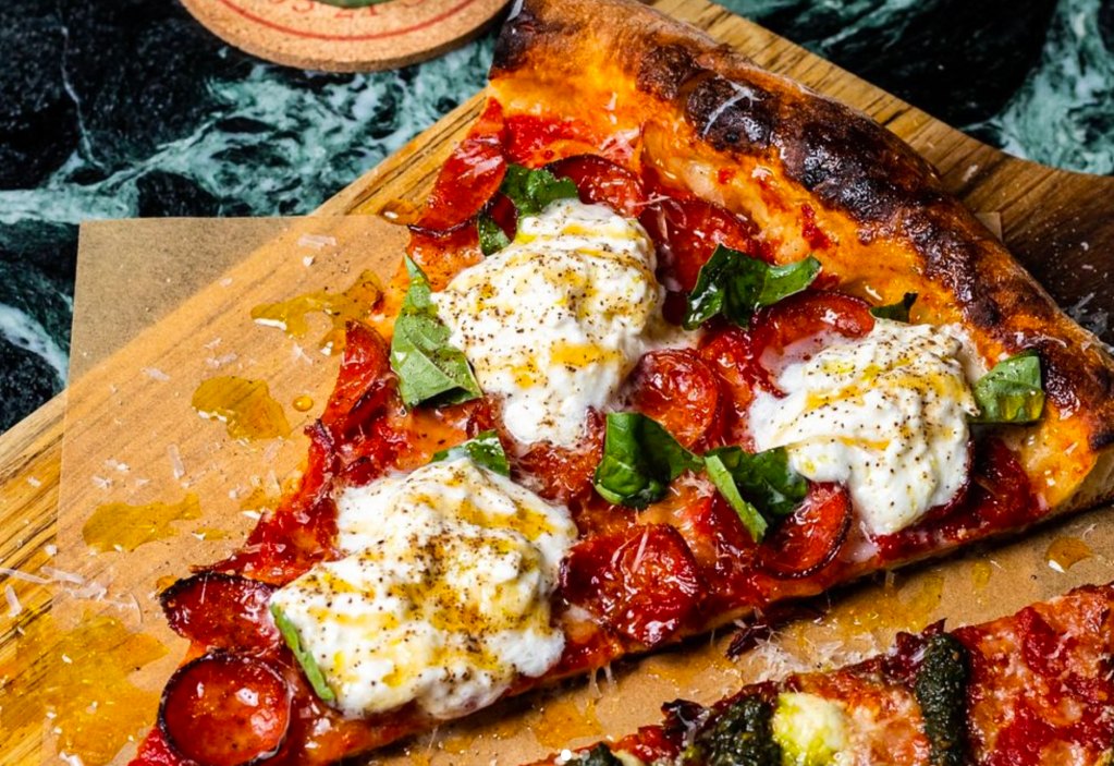 A large pizza slice topped with ricotta, pepperoni, and fresh basil on a wooden board.