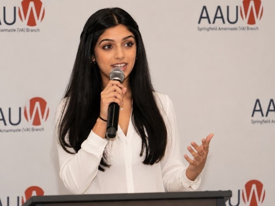 A young woman speaks into a microphone at a podium before a branded backdrop.