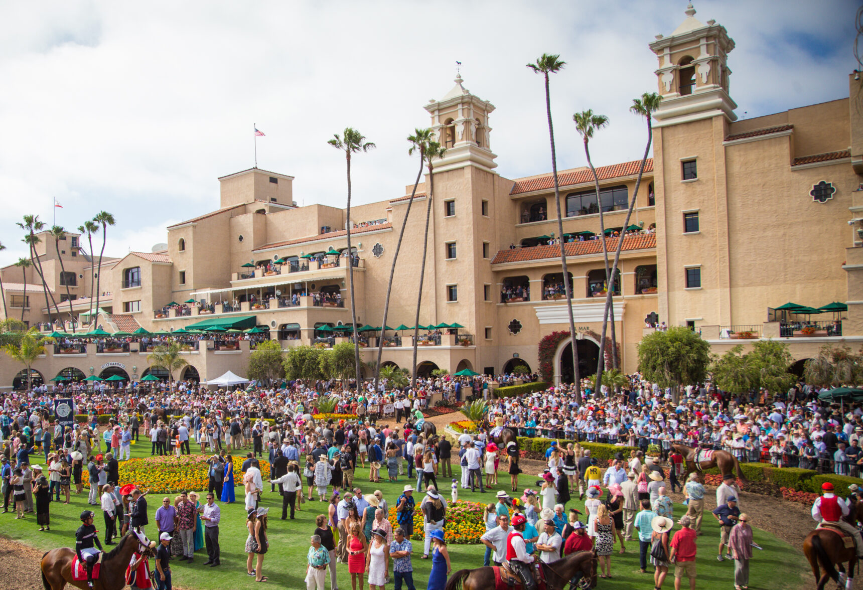 Large crowd at a racetrack with a Spanish Colonial Revival grandstand and tall palm trees.