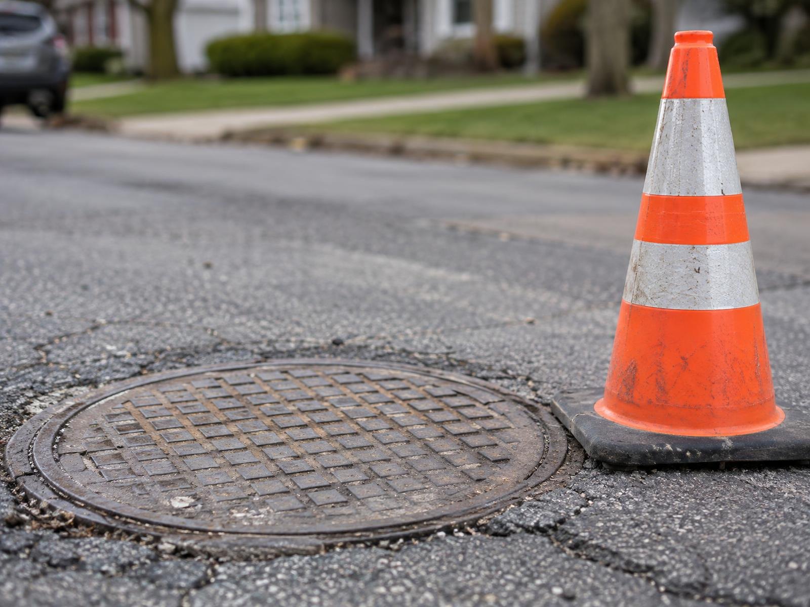A concrete manhole cover on a street with an orange safety cone beside it.