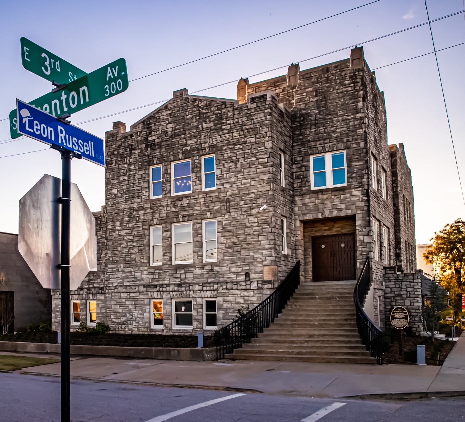 Stone two-story building at a street corner with a Leon Russell street sign at dusk.