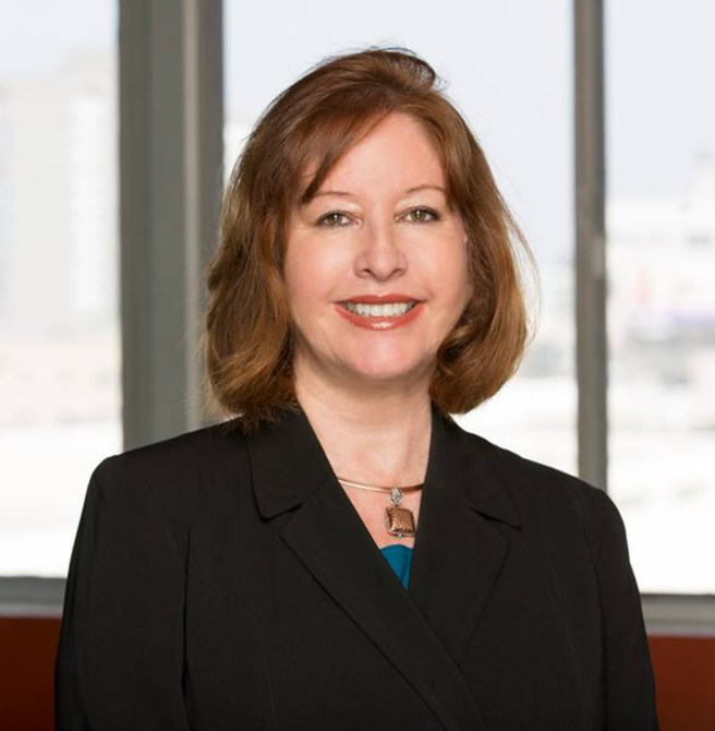 Professional headshot of a smiling woman in a black blazer, standing before office windows.
