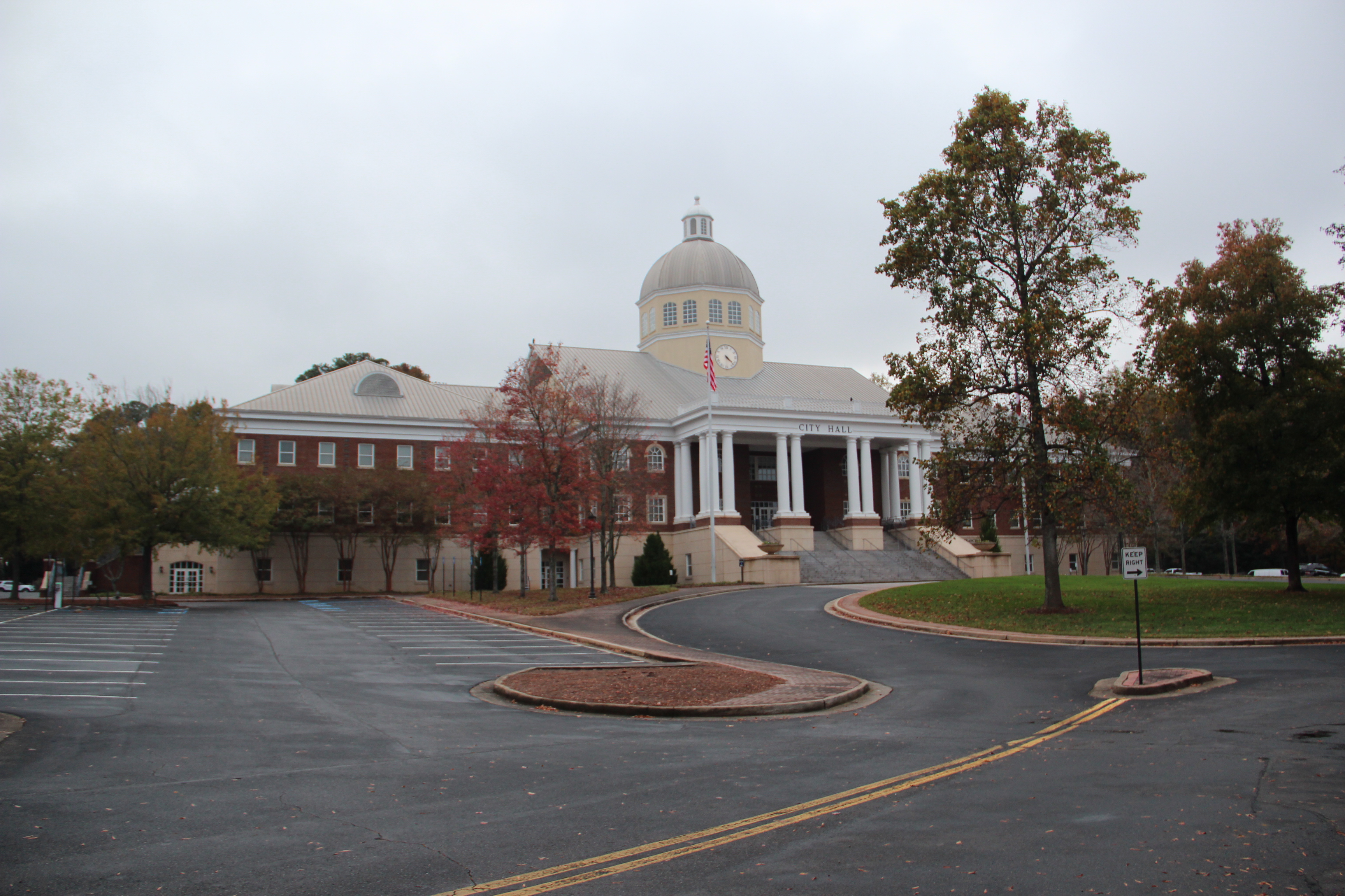 Brick government building with a domed cupola, white columned entrance, and autumn trees under an overcast sky.