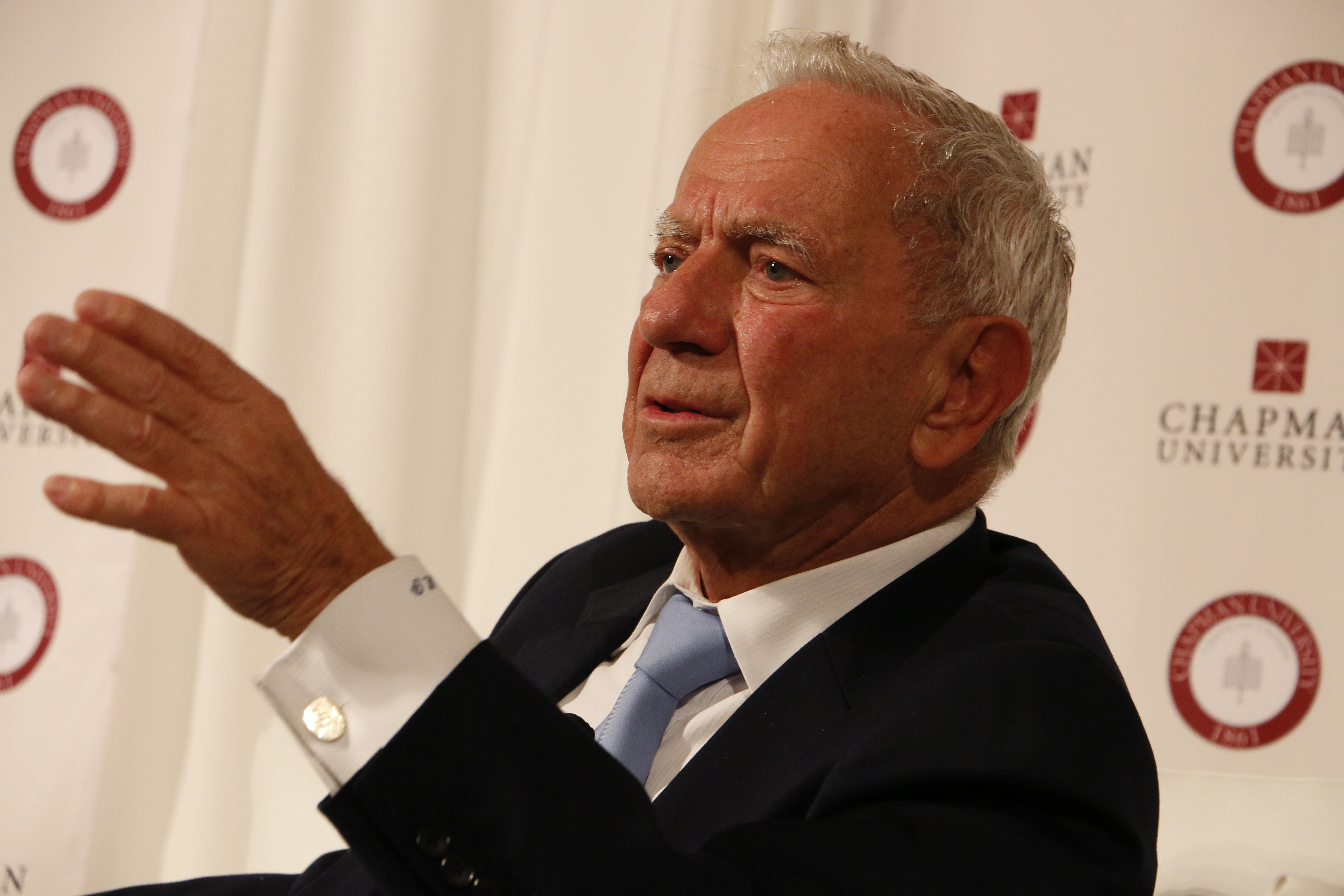 Elderly man in a dark suit gesturing while speaking in front of a university banner.