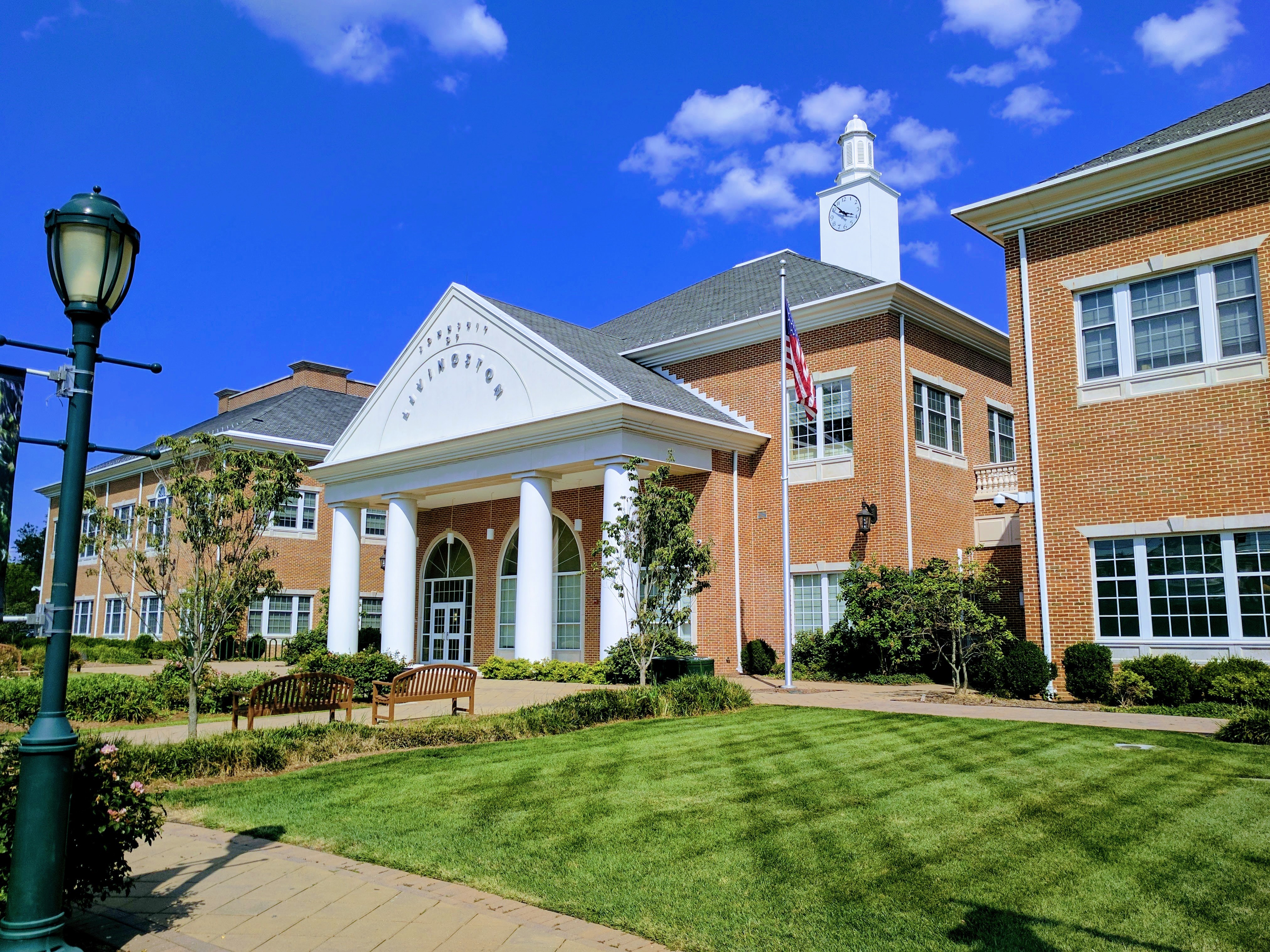 Brick municipal building with white columned portico, clock tower, and American flag on a sunny day.