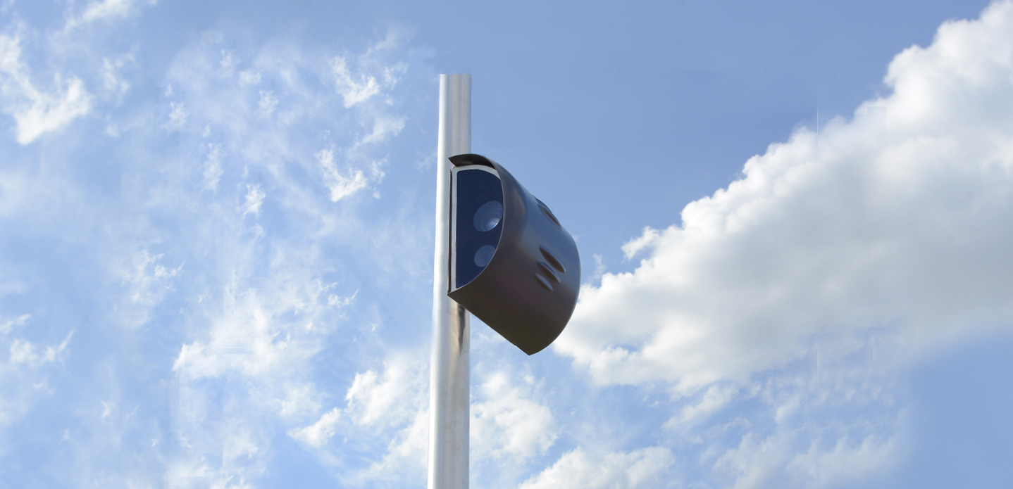 Dark automated traffic enforcement camera mounted on a metal pole against a cloudy sky.