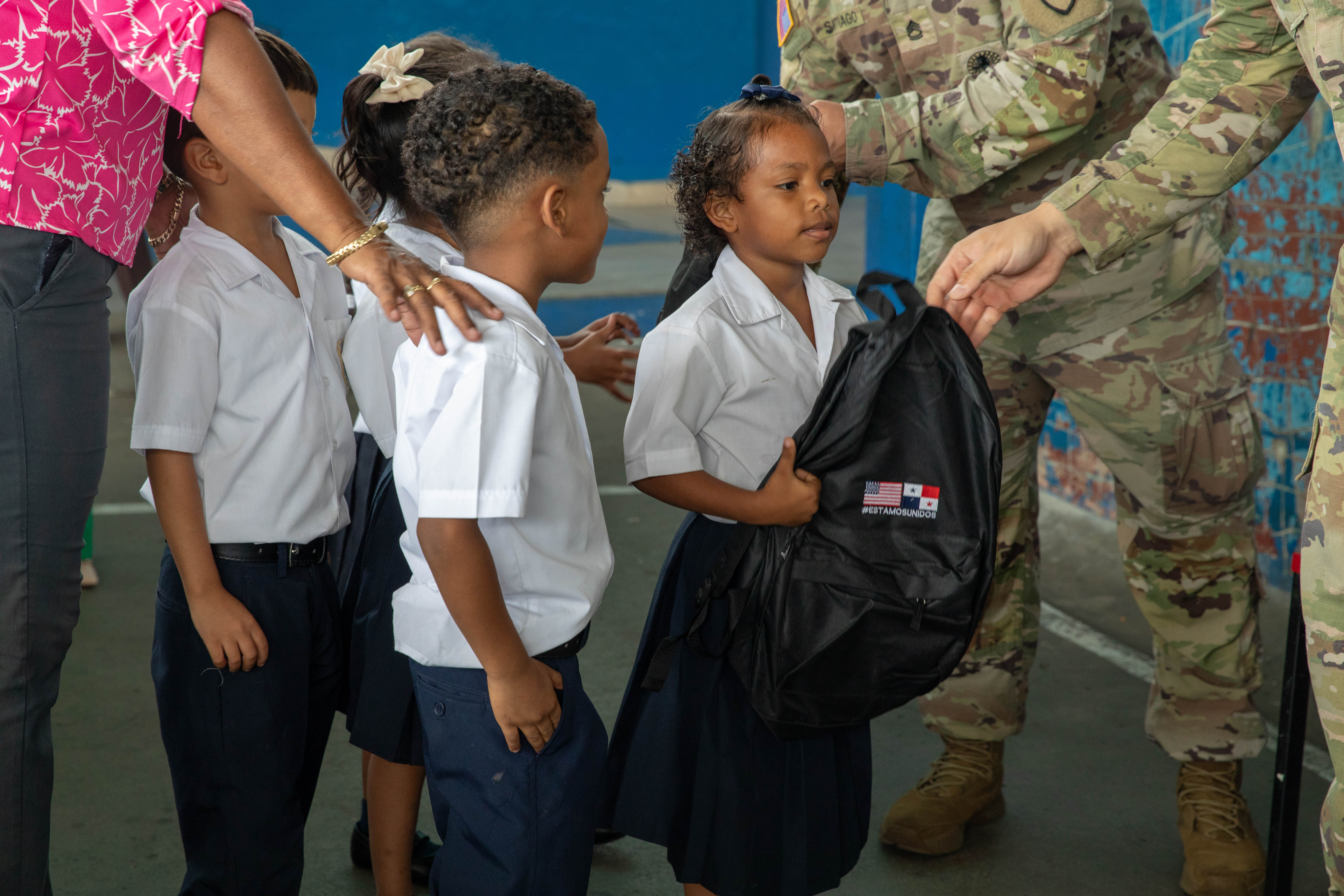 An adult hands a black backpack to a young girl in school uniform at an outdoor event.