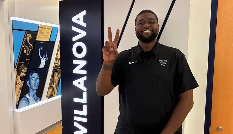 Smiling man in a Villanova Nike polo making a peace sign in front of a Villanova Athletics banner.