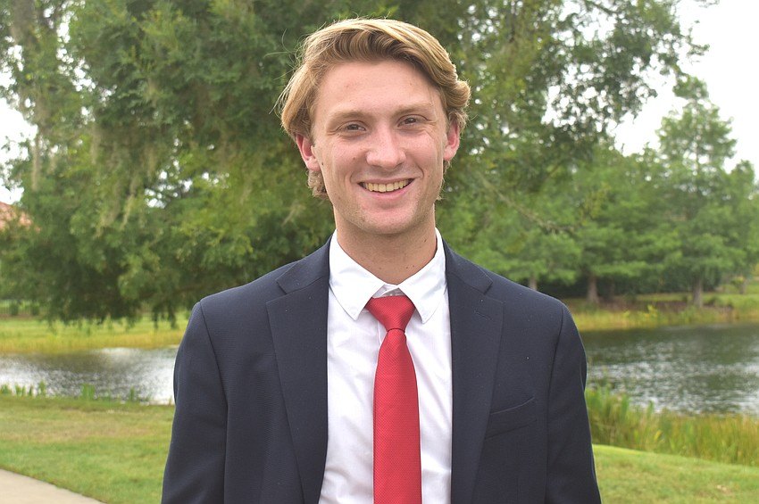 A young man in a navy suit and red tie posing outdoors near a pond.