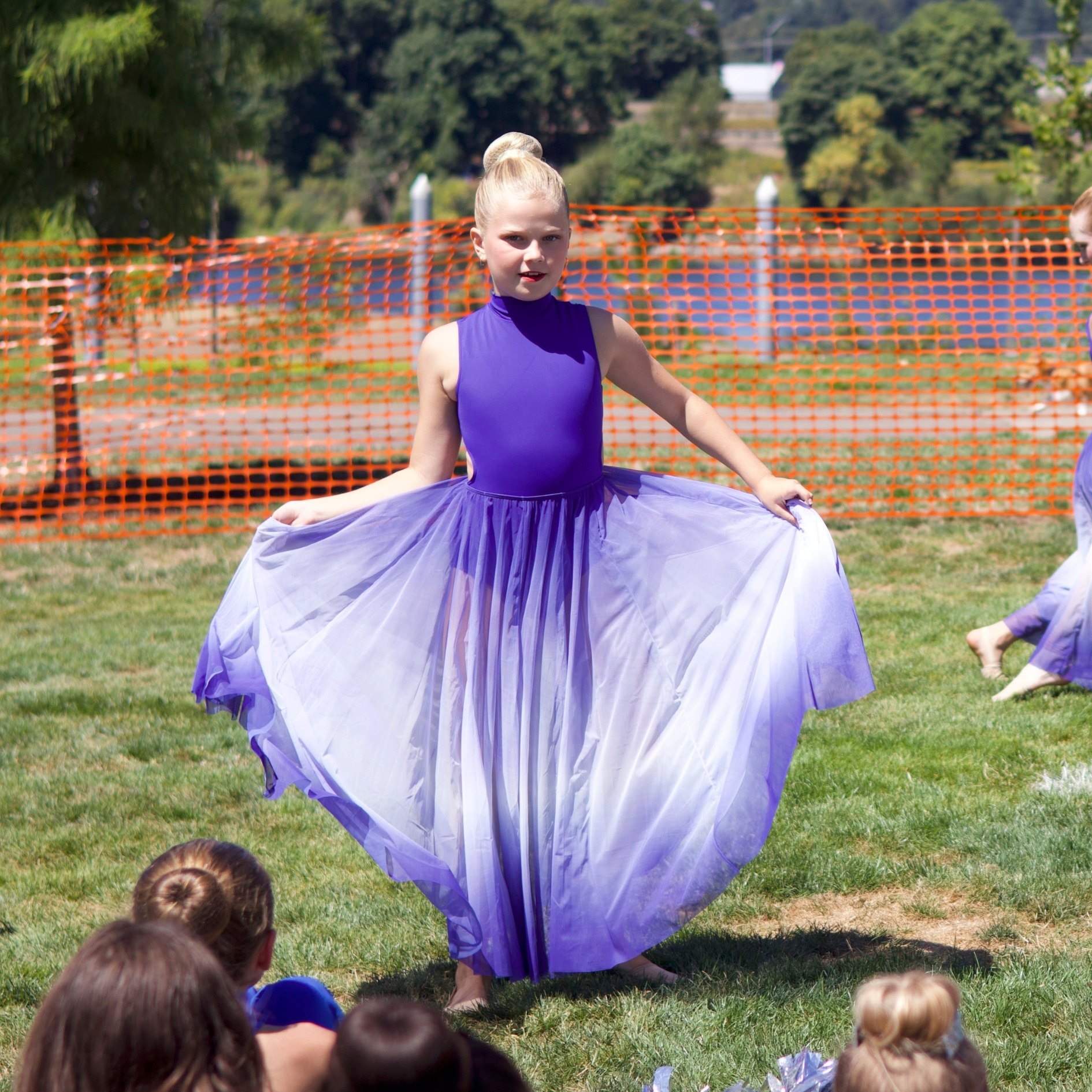 A young girl in a purple dance costume twirls on a grass field outdoors.