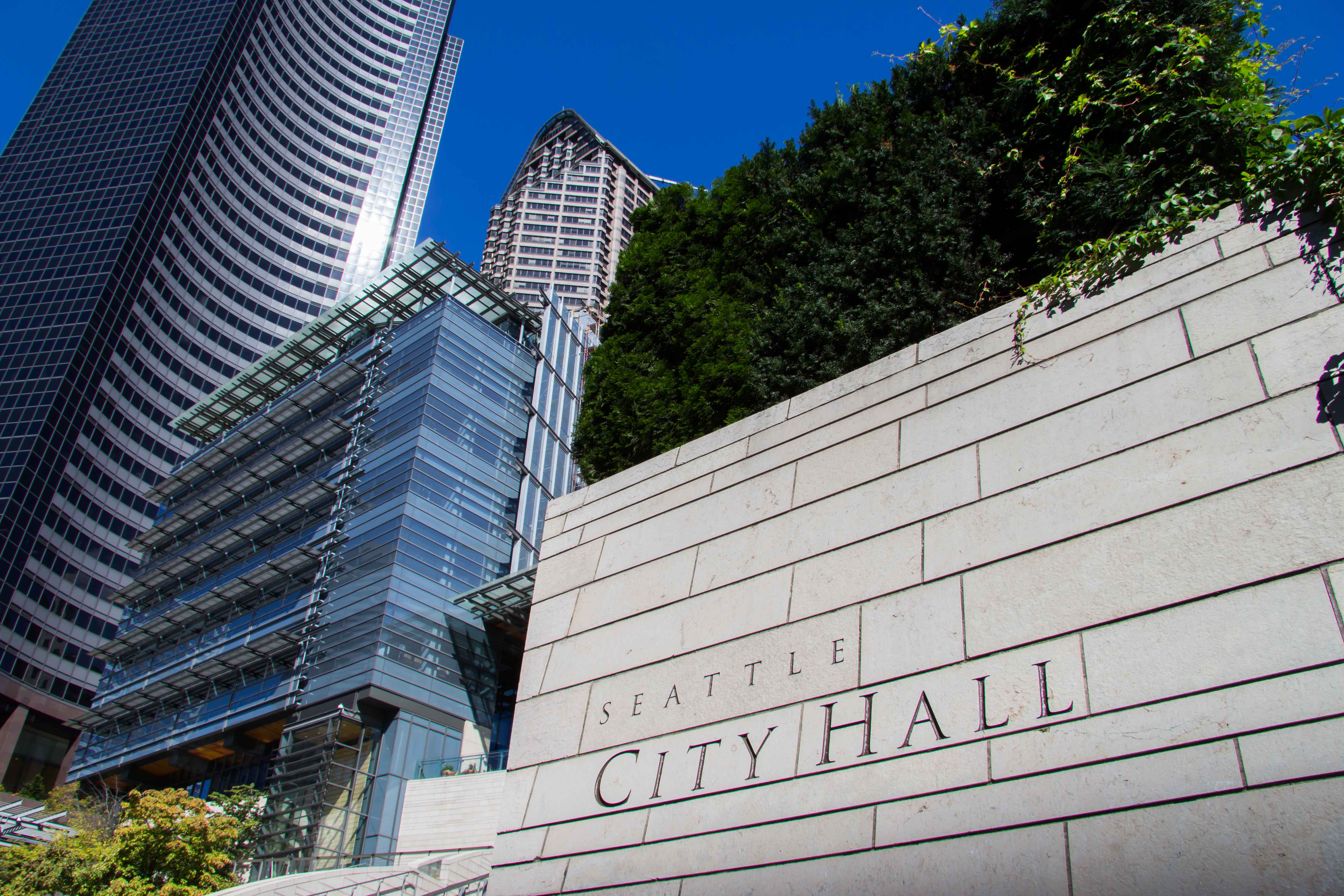 Stone sign reading 'Seattle City Hall' with glass skyscrapers and trees in the background.