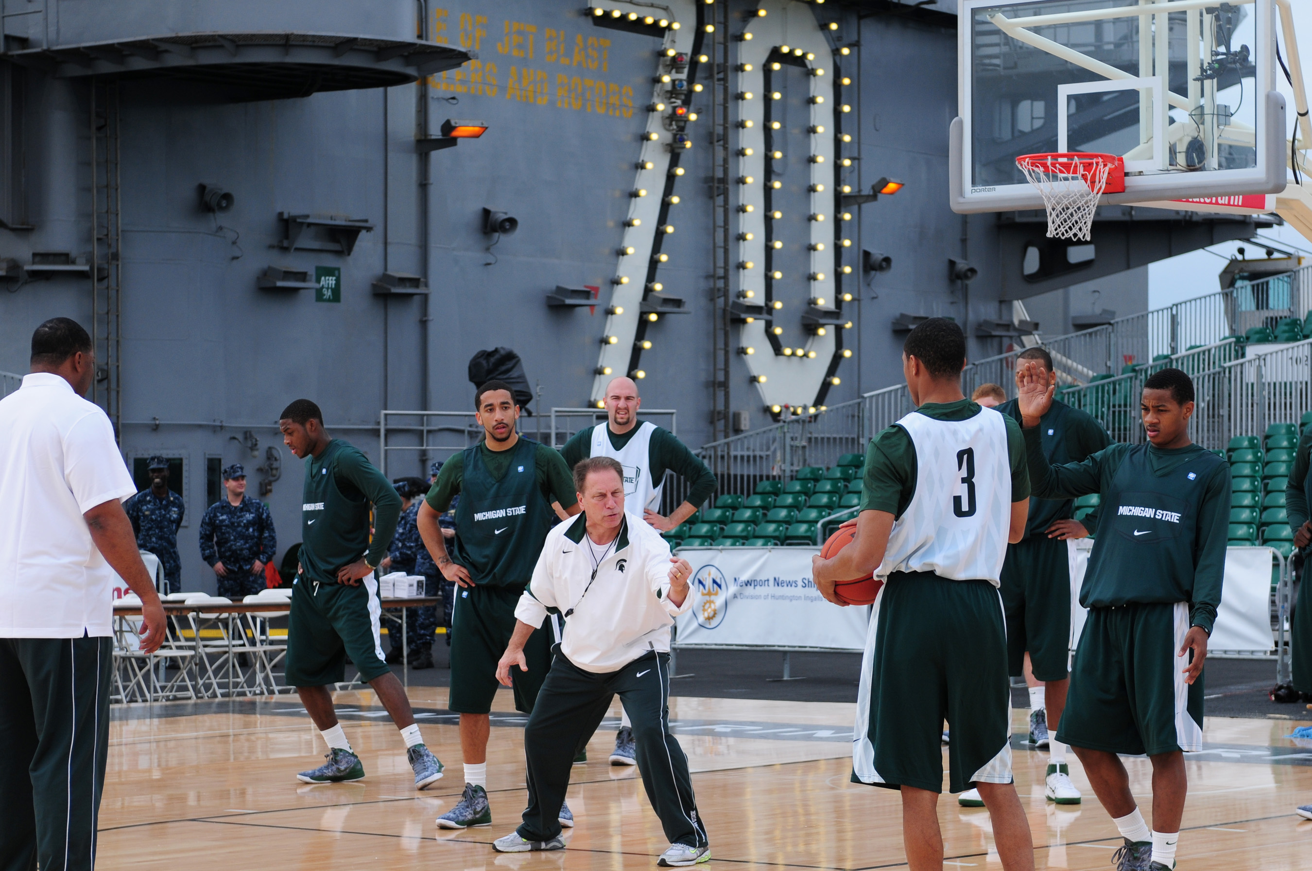 A coach in a white shirt instructs players in green Michigan State uniforms on a basketball court outdoors.