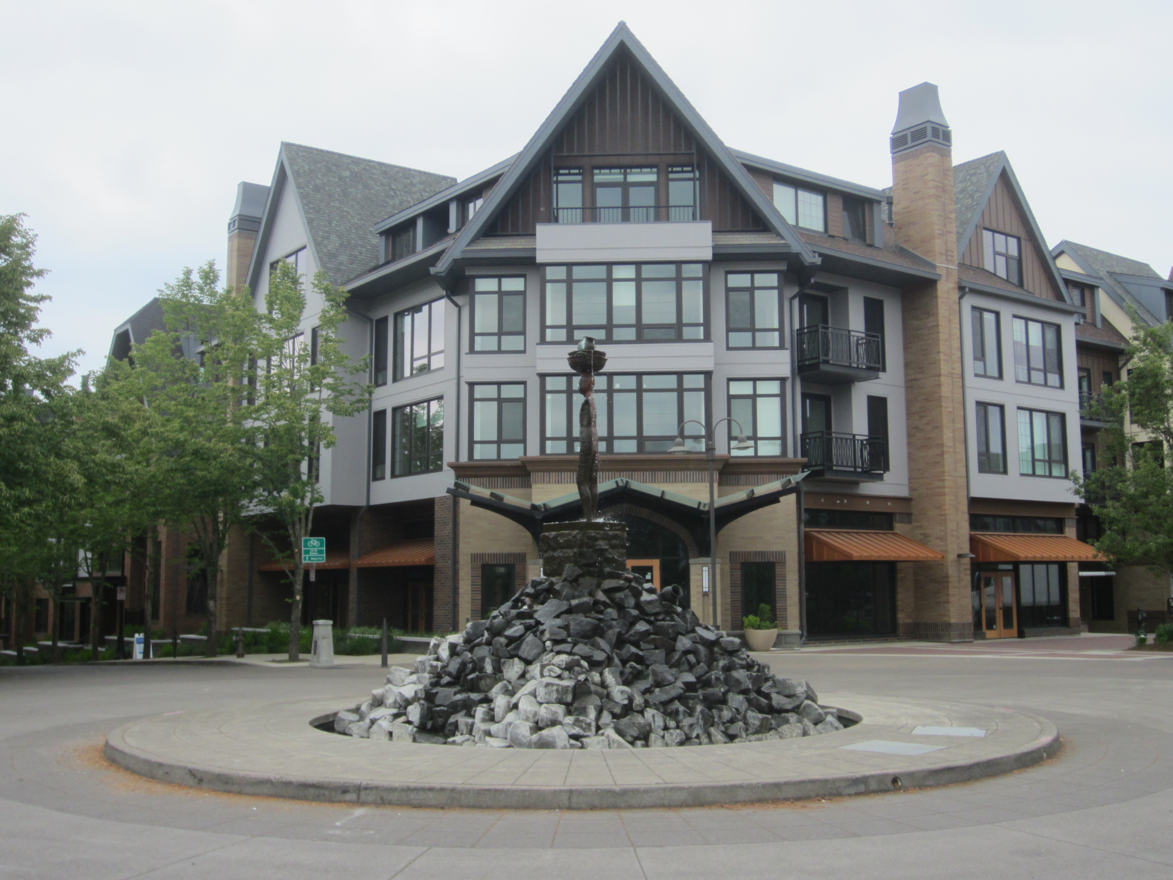 Multi-story building behind a circular plaza with a tall rock sculpture and trees.