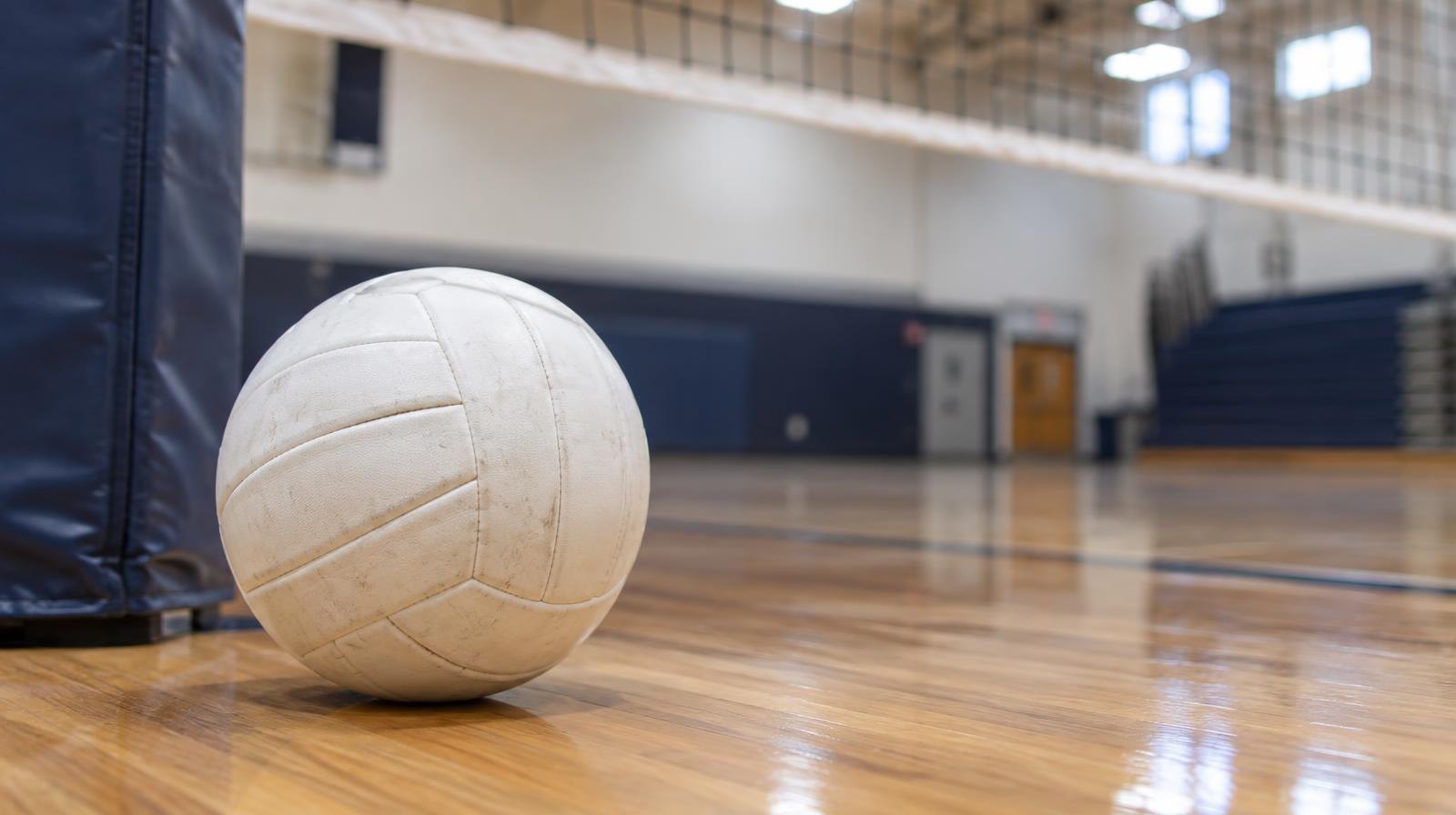 A white volleyball resting on a hardwood gym floor beside a net post.