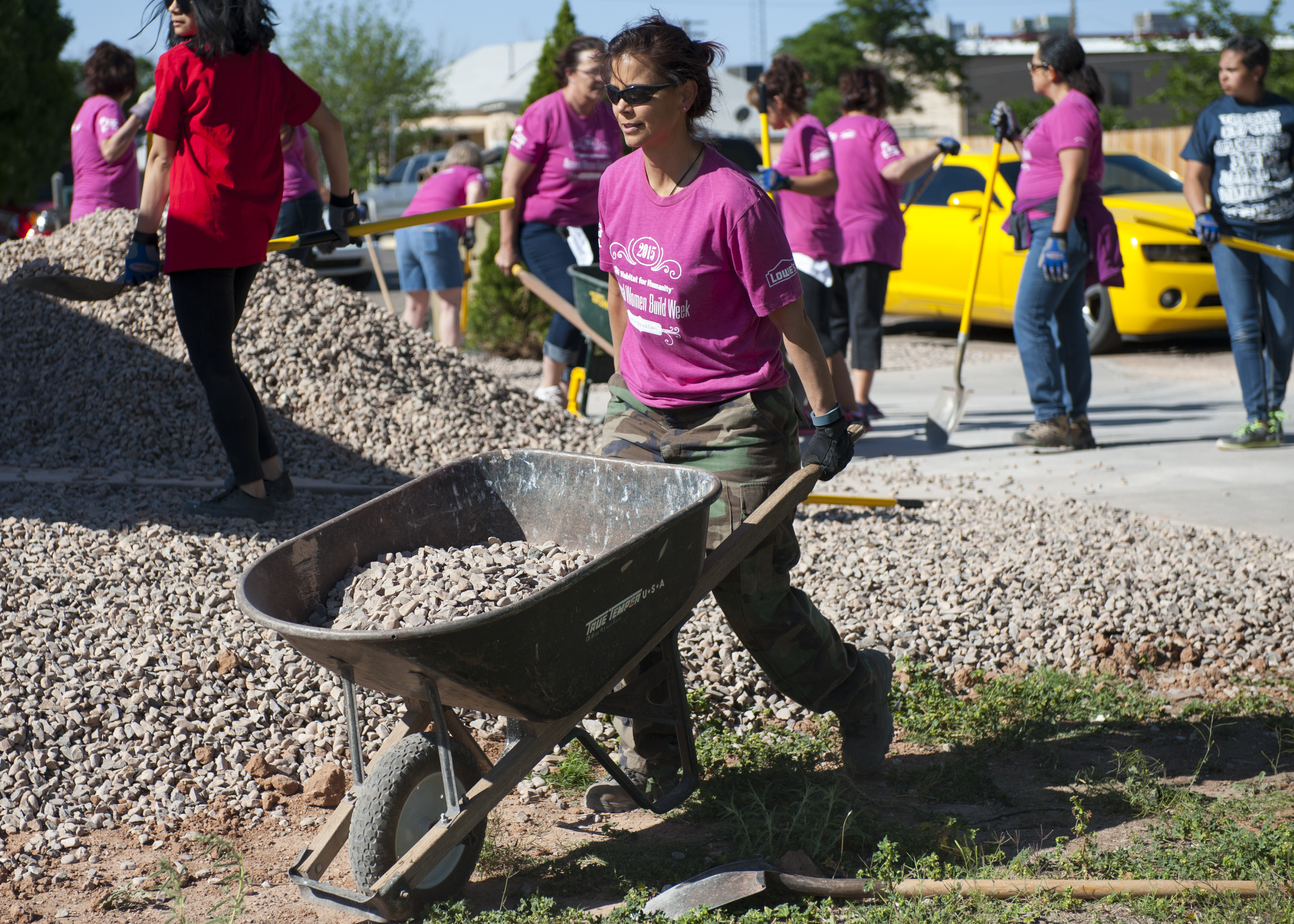 Volunteers in matching pink shirts use shovels and a wheelbarrow at a Habitat for Humanity site.