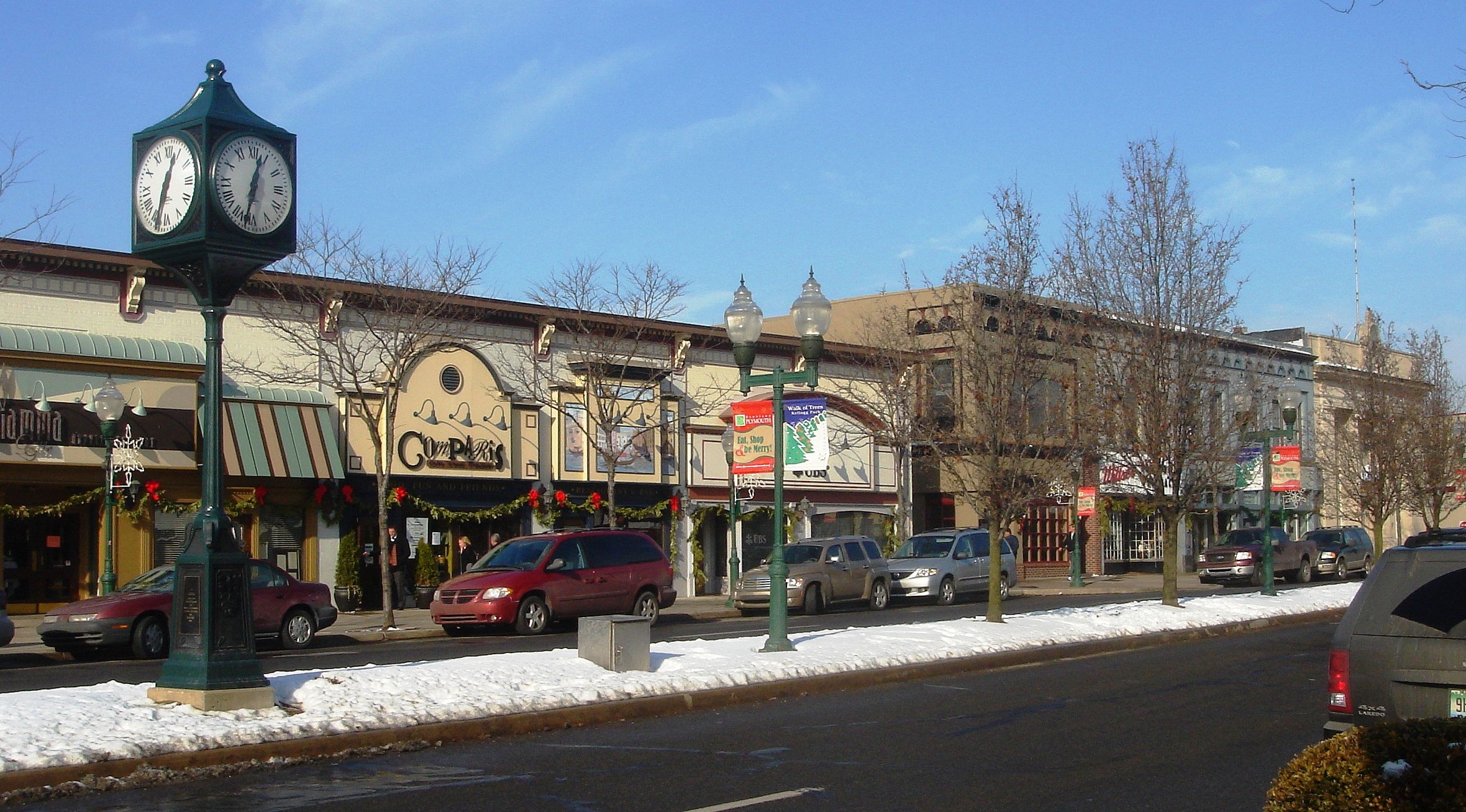 Winter street scene in downtown Plymouth with a four-faced clock, storefronts, and light snow.
