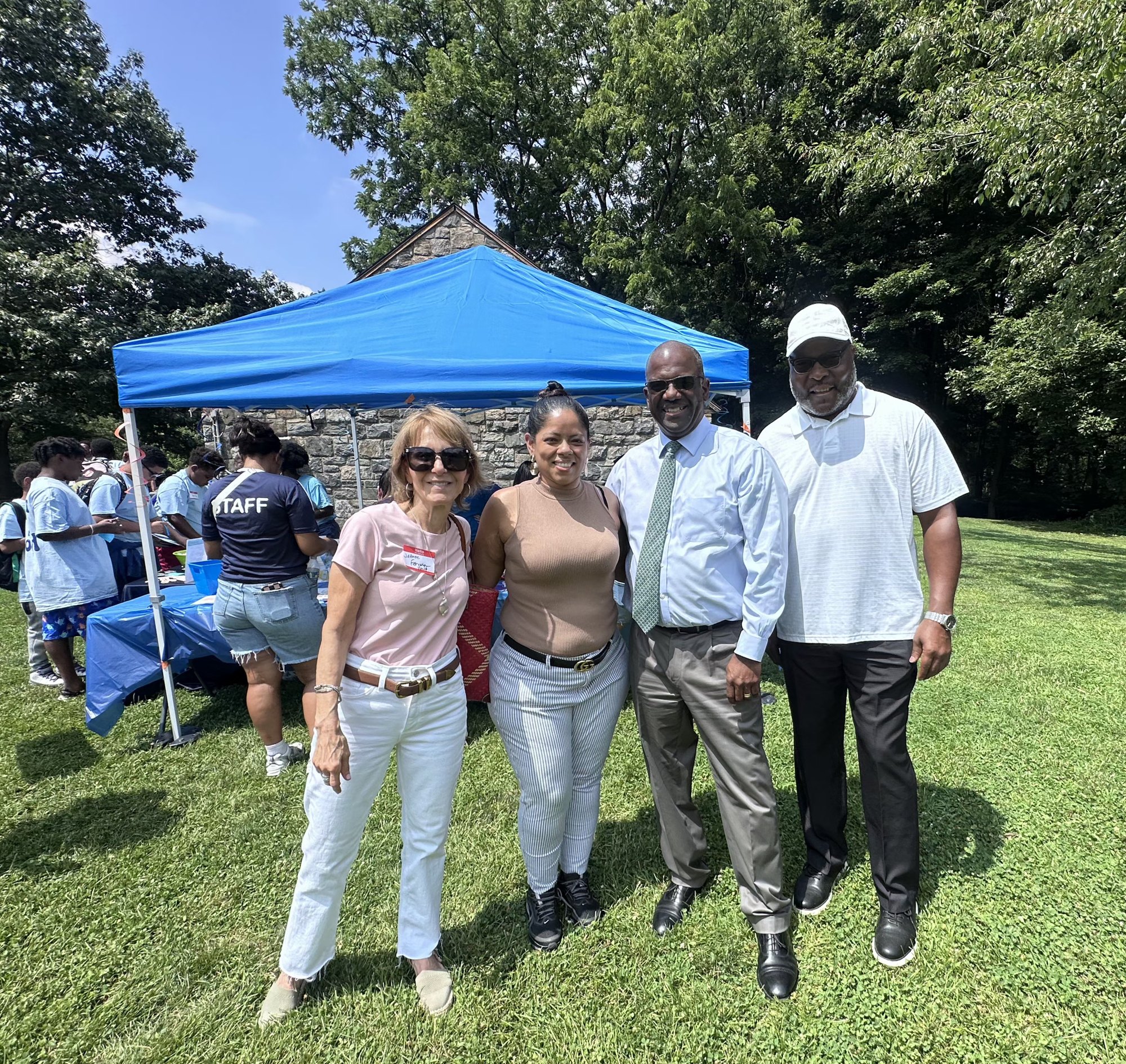 Four people posing together outdoors at a community event with a blue tent and trees in the background.