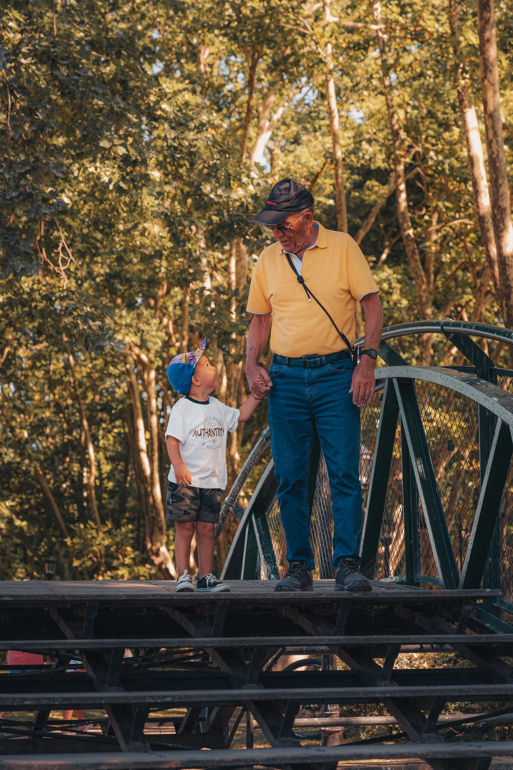 A grandfather and grandson enjoying a sunny day walk on a rustic bridge surrounded by trees.