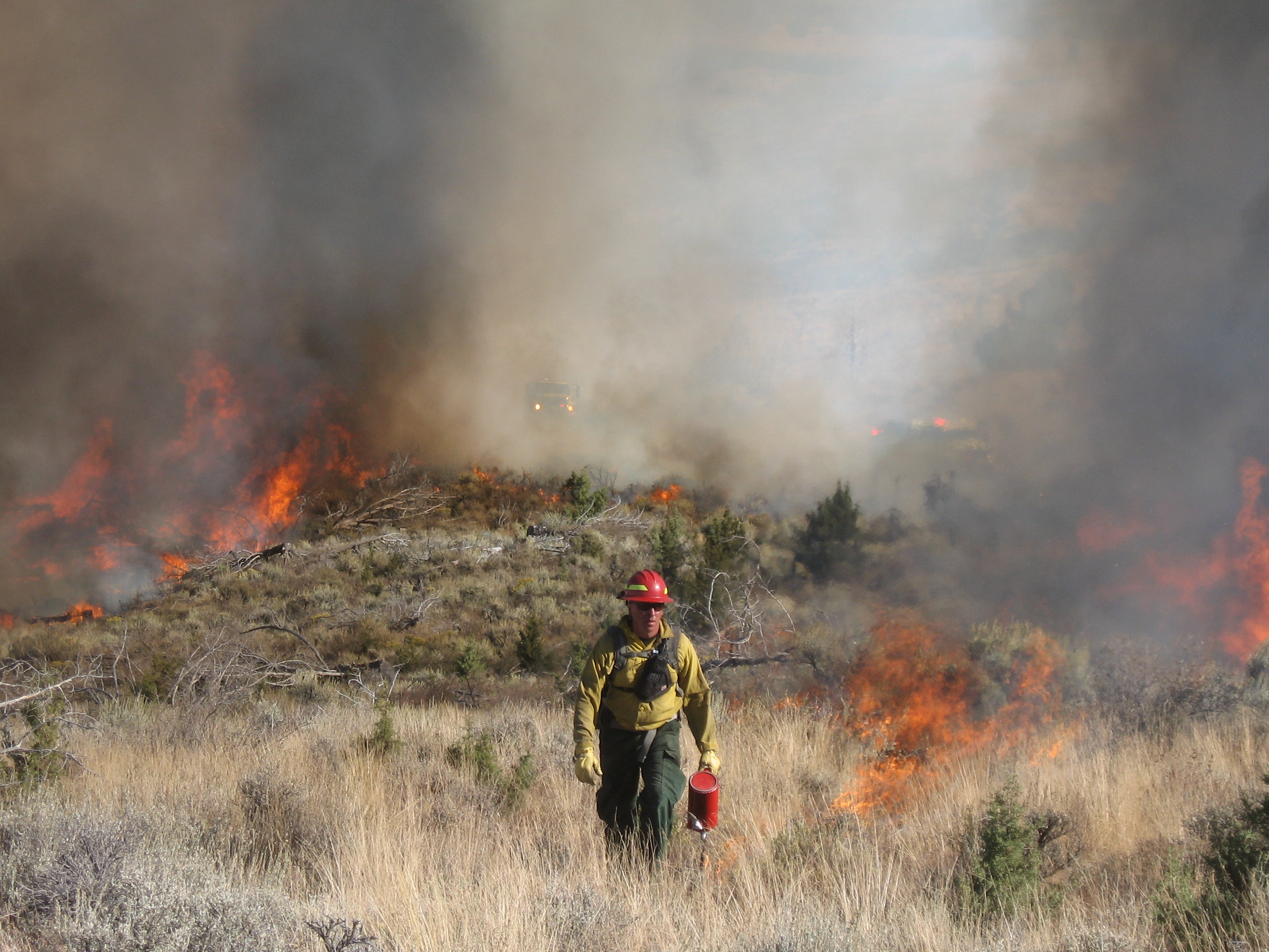 A firefighter in yellow gear carries drip torches near an active brush fire with heavy smoke.