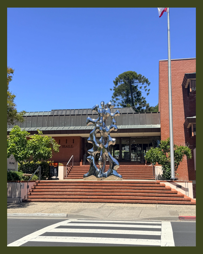 Large abstract metal sculpture with human figures at the steps of a brick civic building.