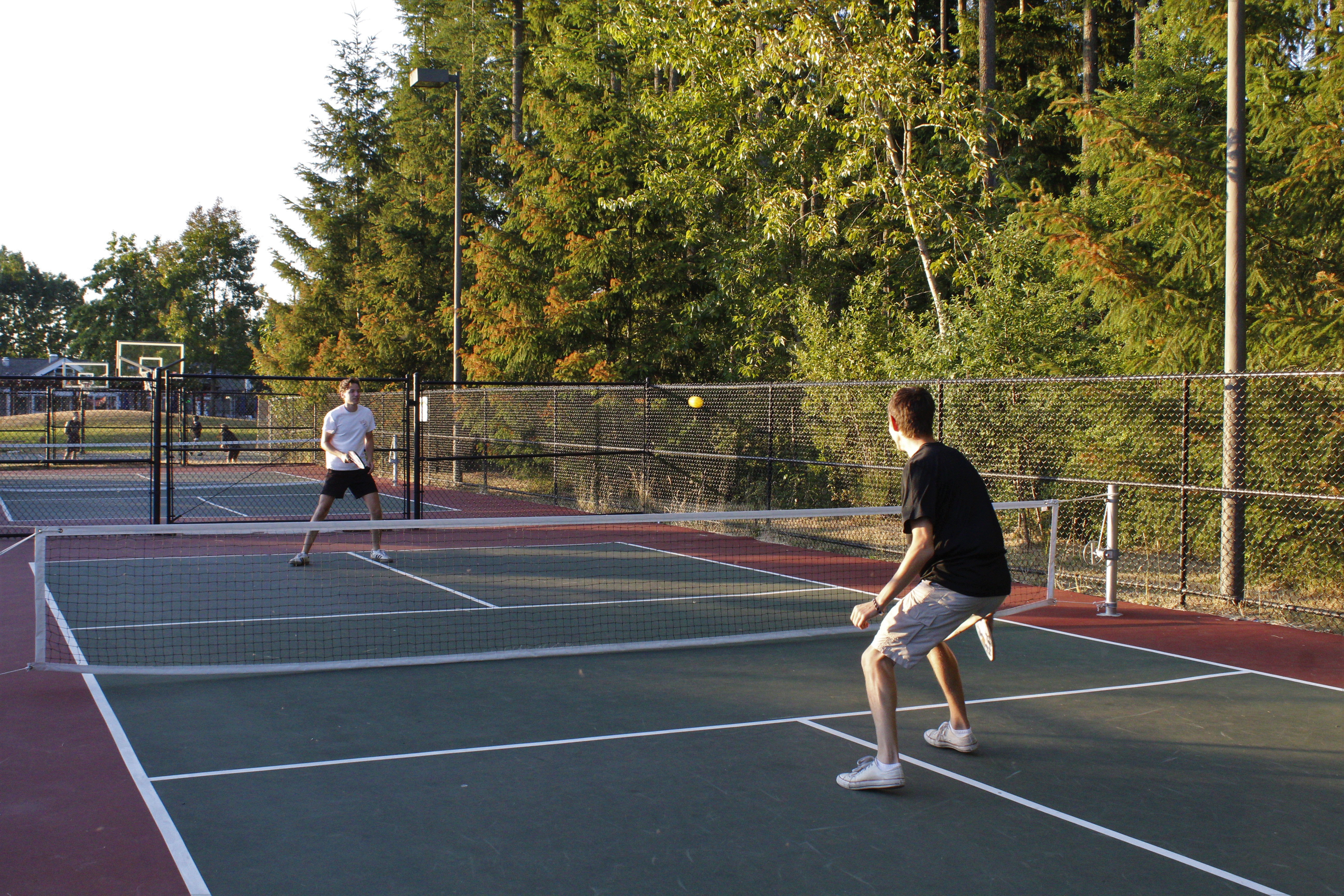 Two people playing pickleball on an outdoor court, ball visible mid-air.