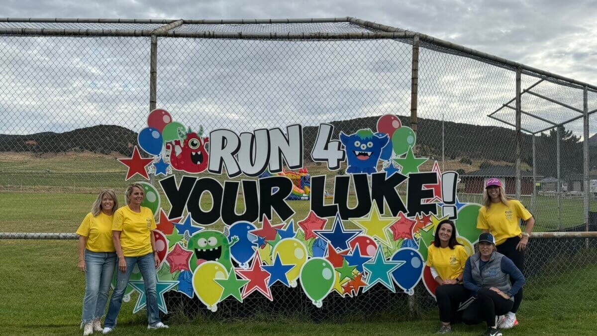 Five people in yellow shirts pose in front of a colorful Run 4 Your Luke banner at an outdoor event.