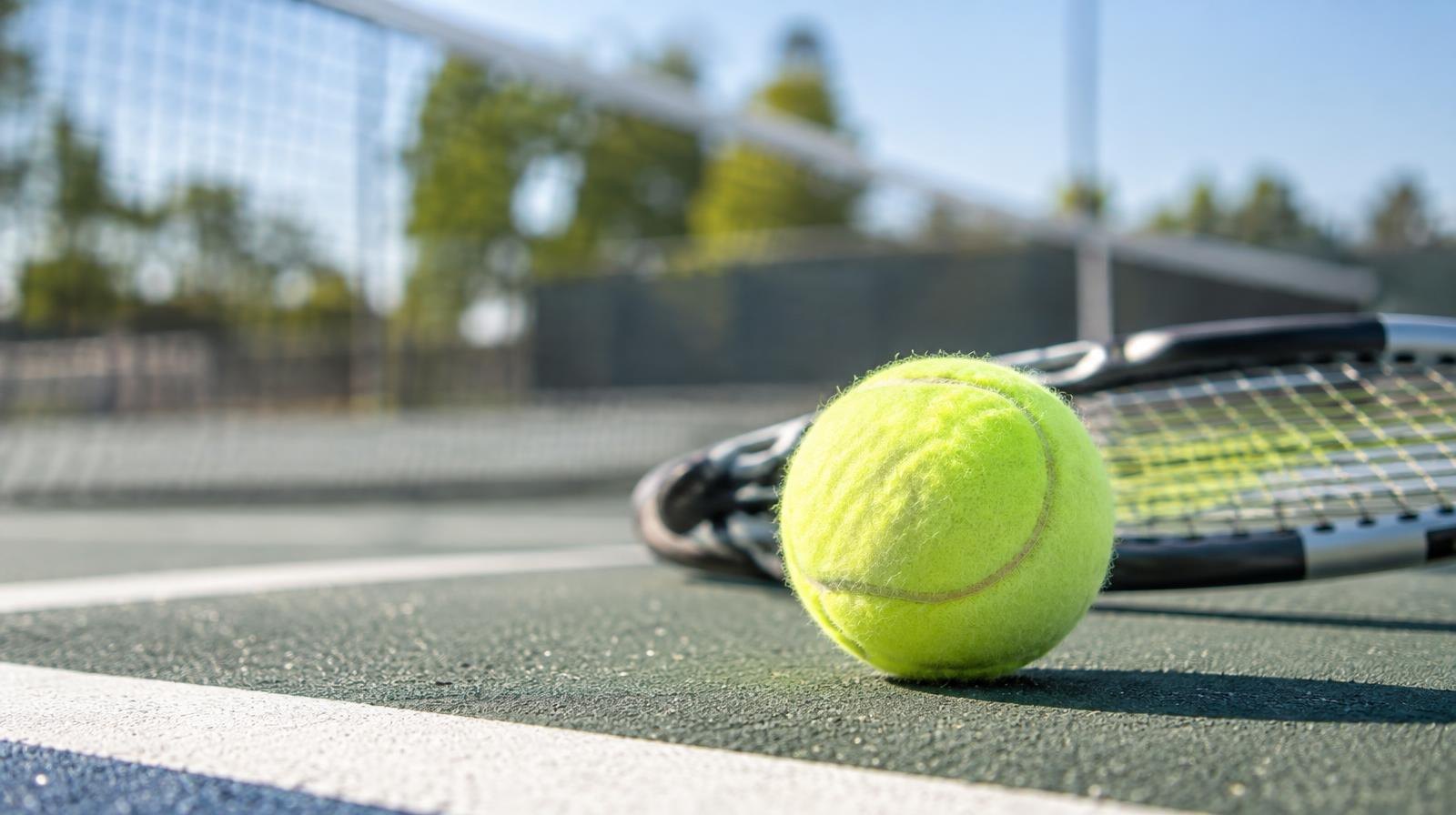 A yellow tennis ball beside a racket on a hard court with a net in the background.