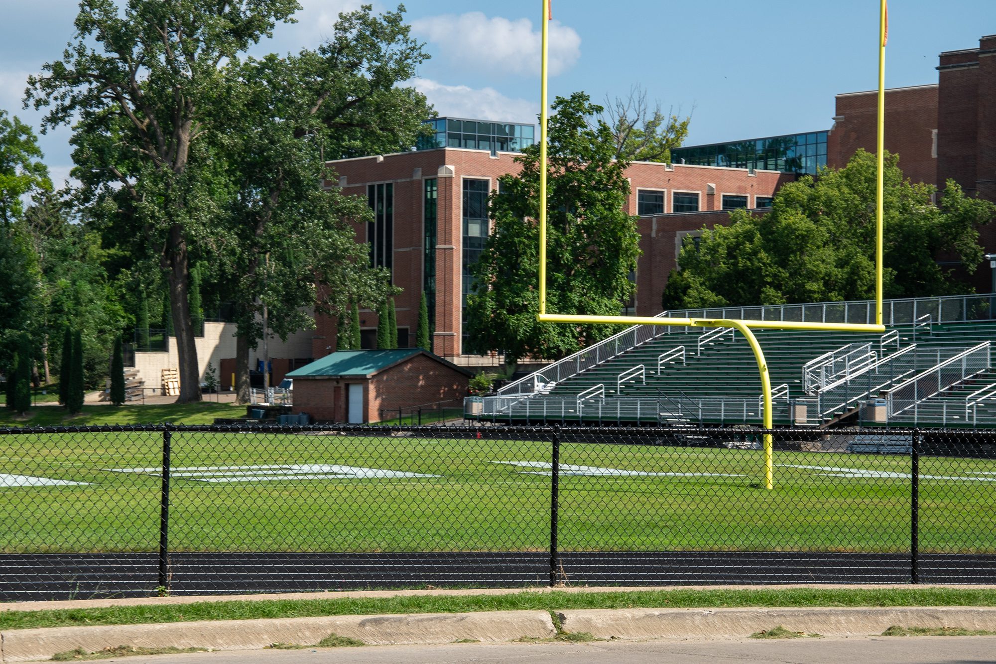 Football field with yellow goalpost, bleachers, and brick school building in background.