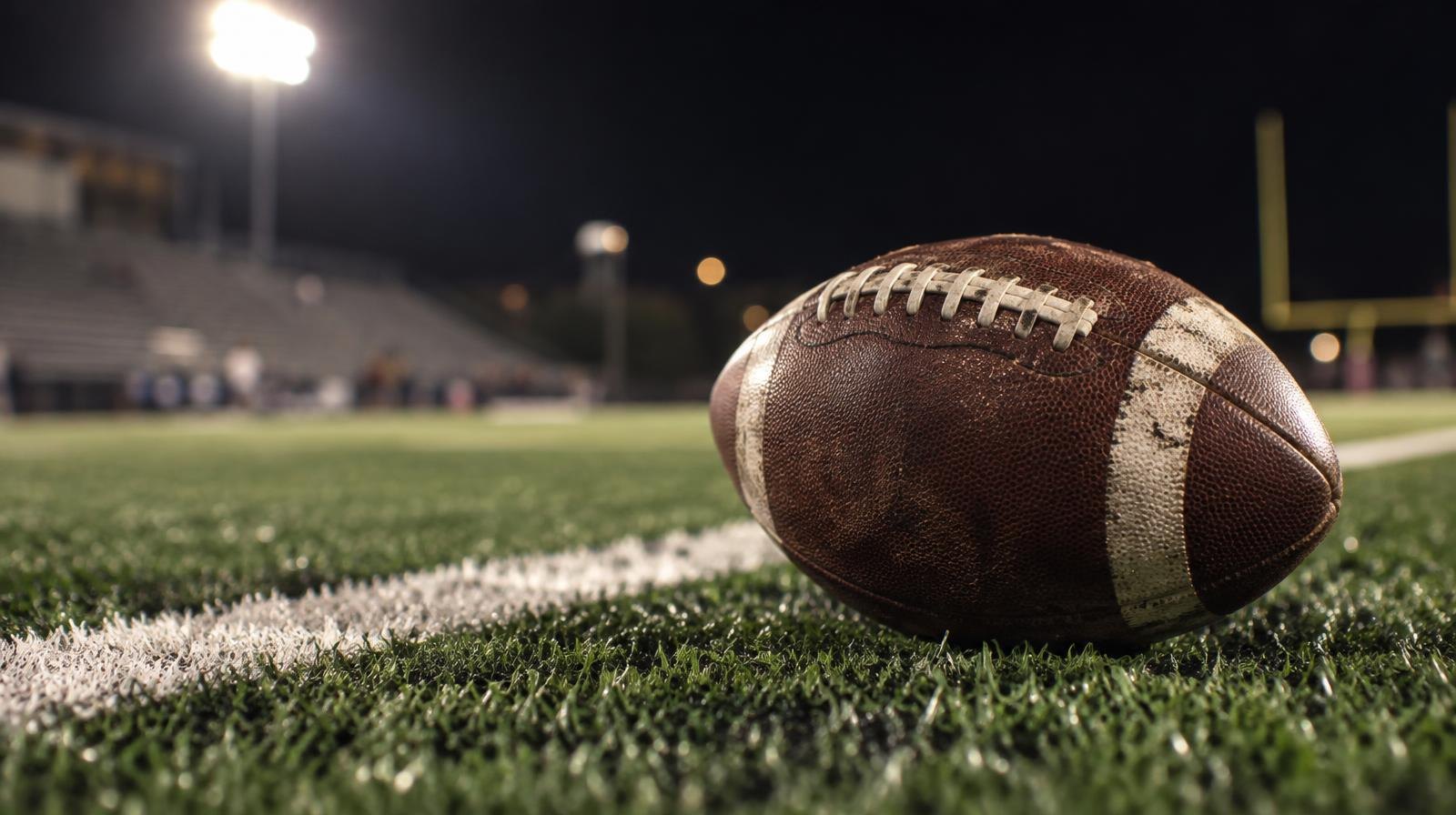An American football on green artificial turf beside a white yard line under stadium lights.