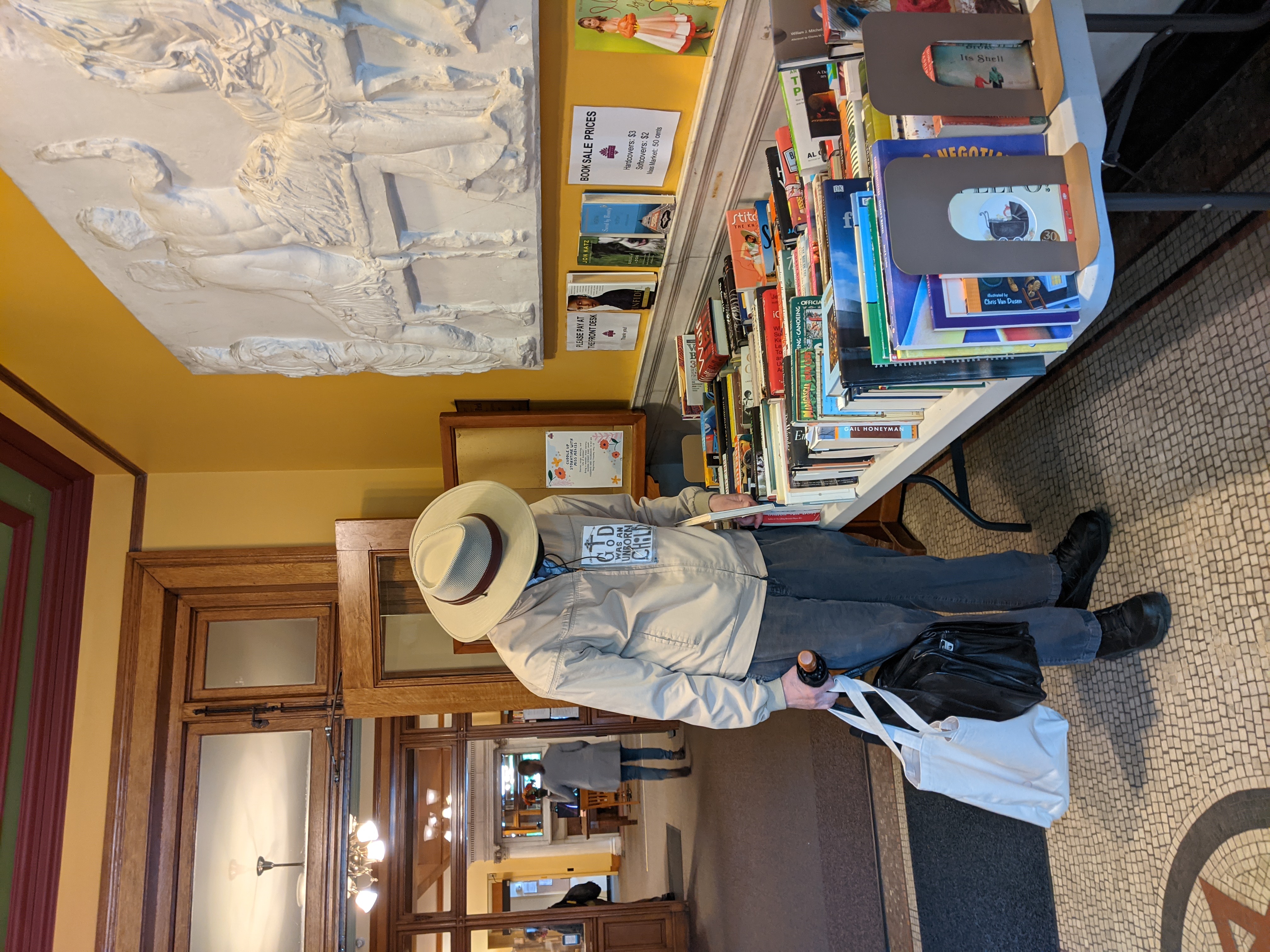 A person in a wide-brimmed hat browses books spread across a table at an indoor book sale.