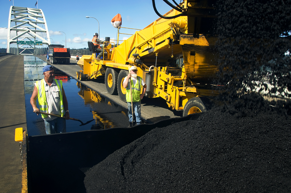 Two workers in yellow safety vests stand near a large yellow asphalt paving machine on a road.