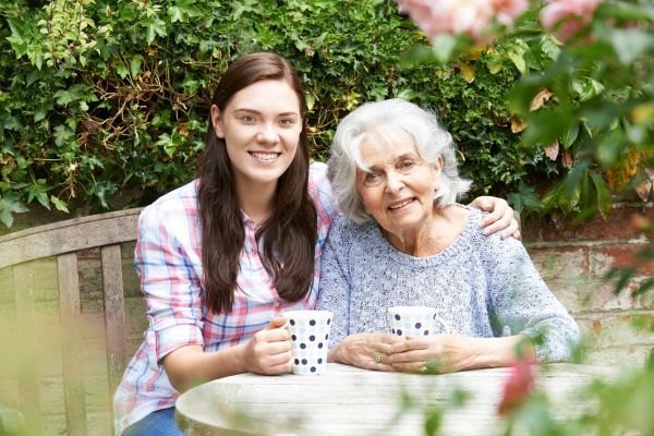 A younger woman and an elderly woman sit together outdoors, smiling and holding mugs.