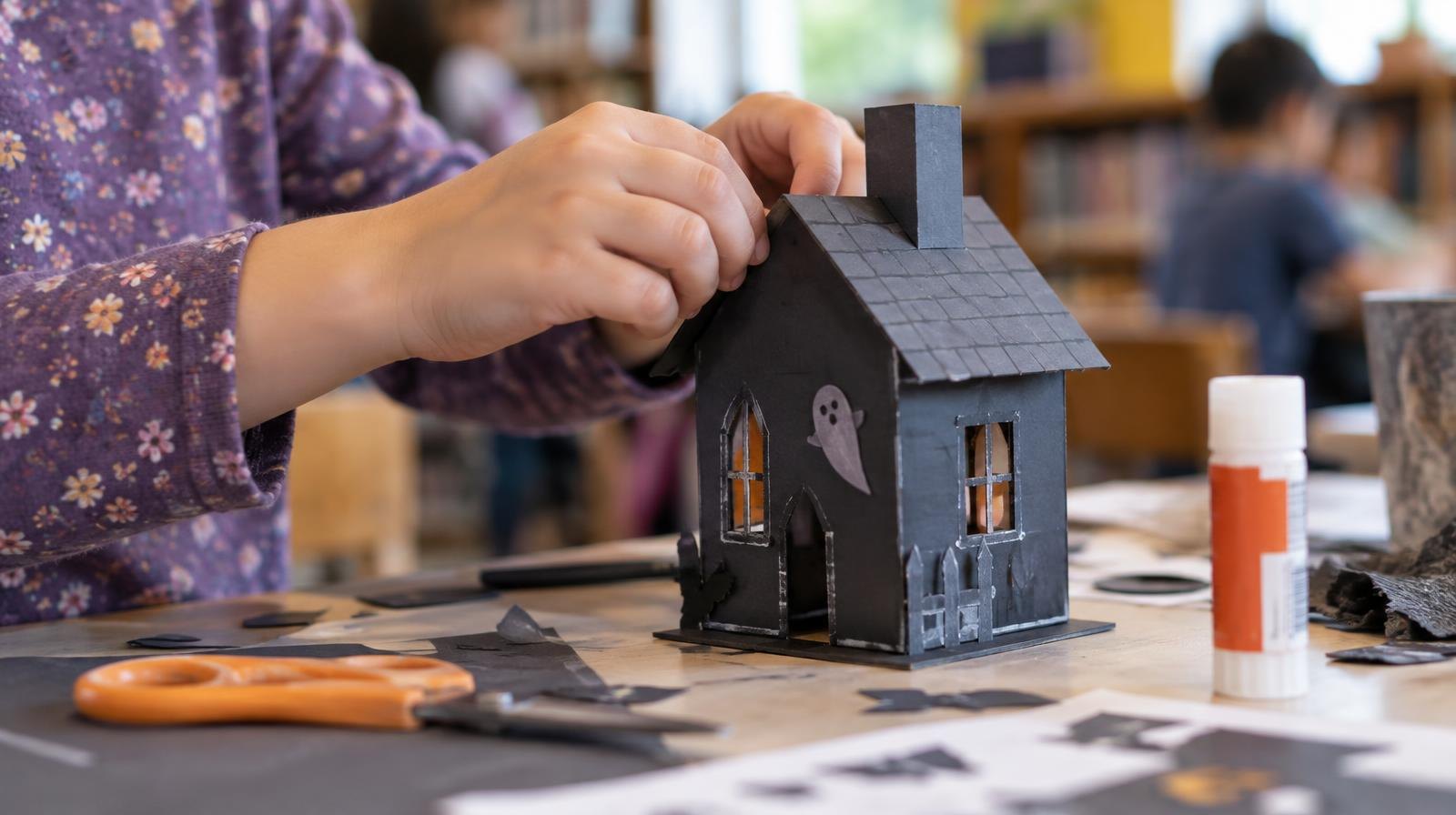 Hands folding and gluing paper into a small haunted house model on a table.