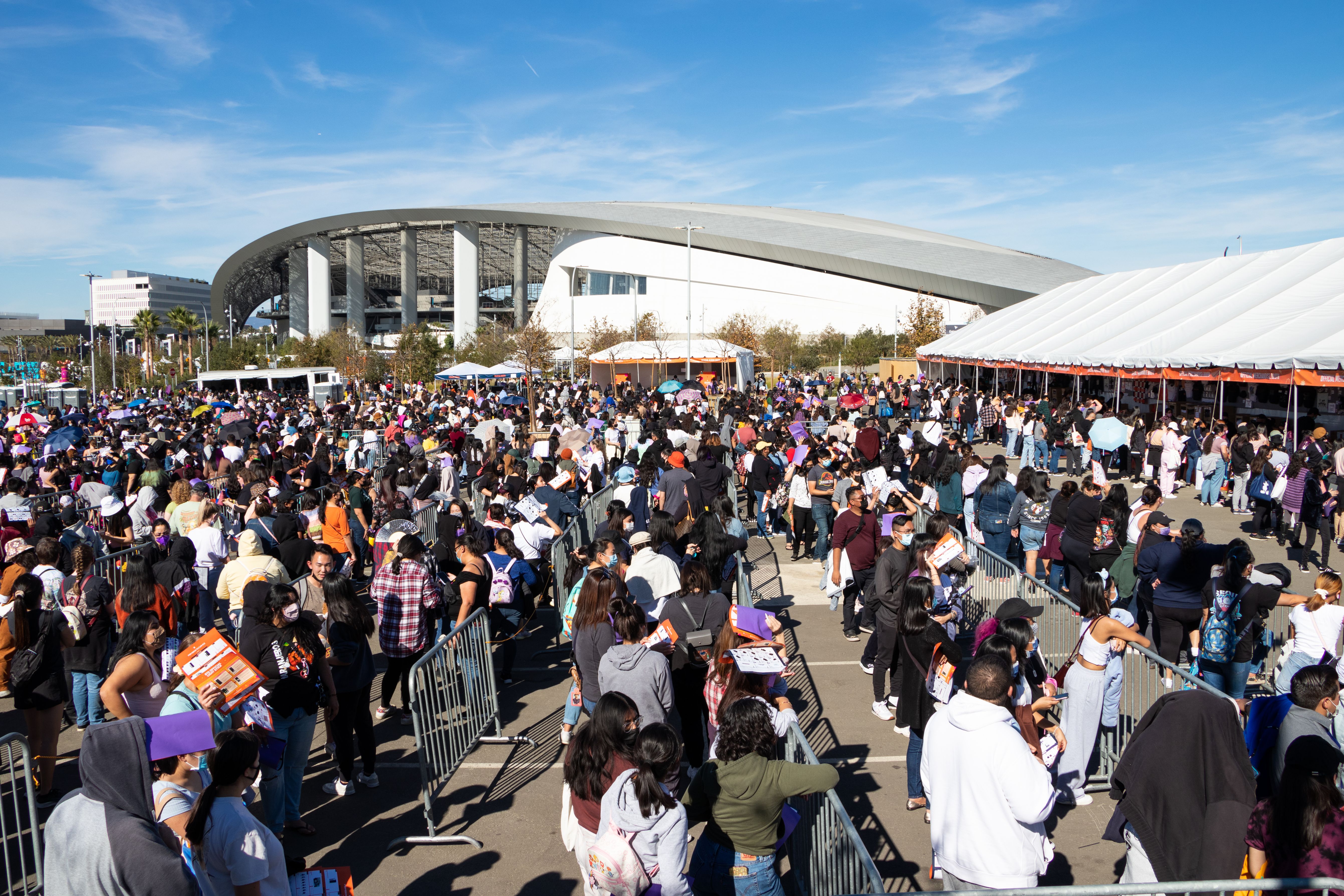 Large crowd gathered in a parking lot outside SoFi Stadium under a blue sky.
