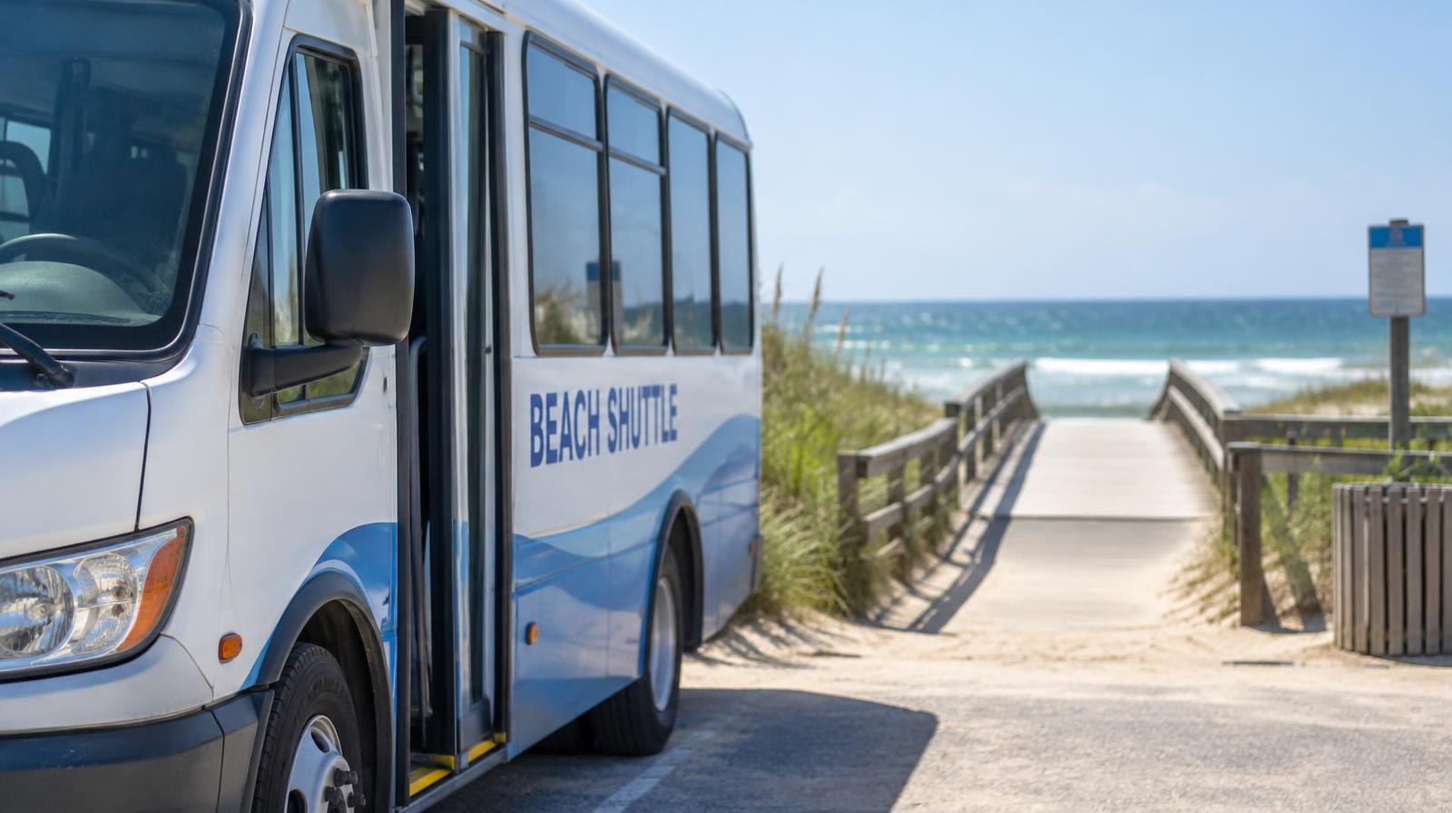 Small transit shuttle bus parked beside a sandy beach boardwalk on a sunny day.