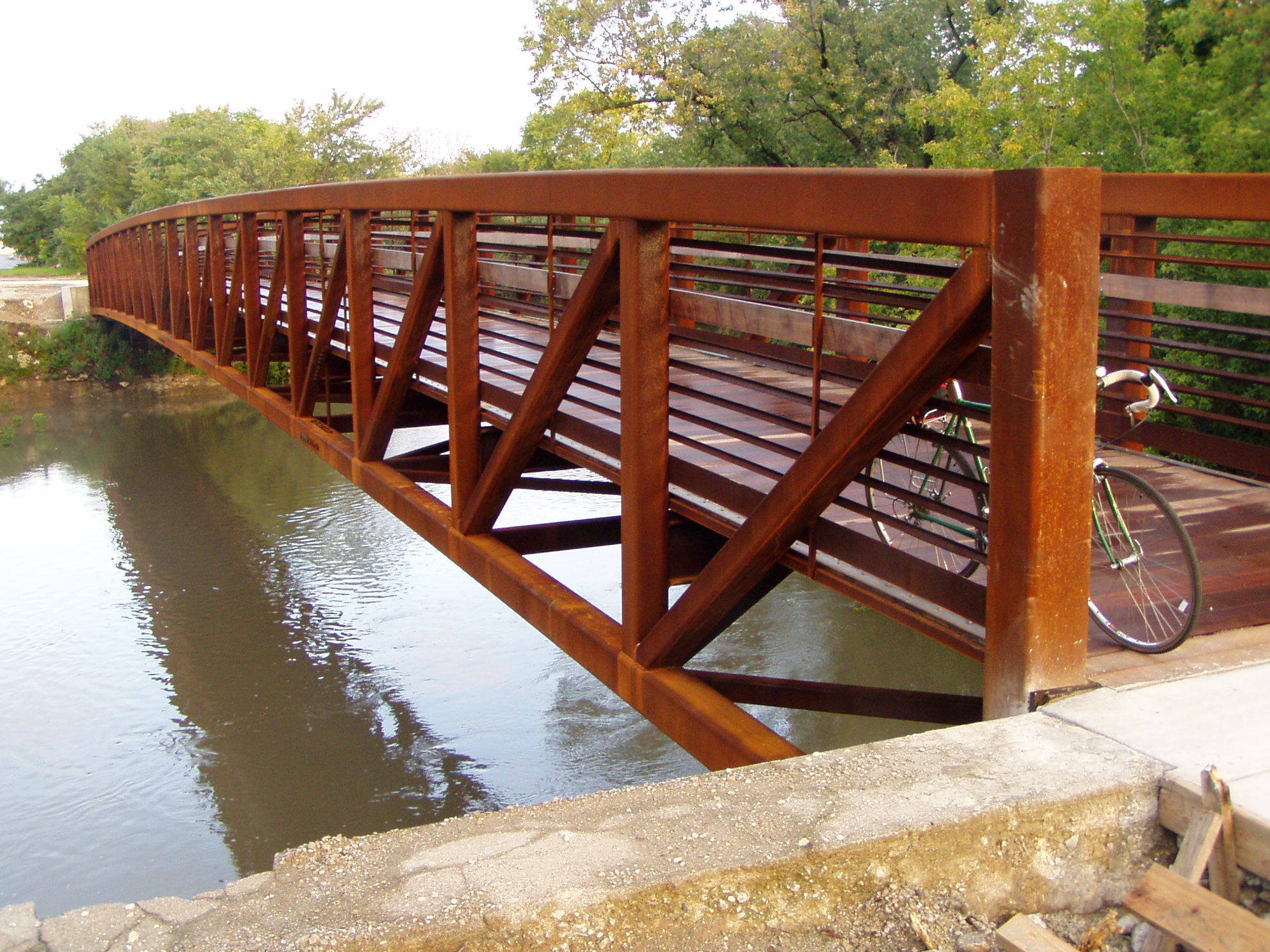 A rust-colored steel truss pedestrian bridge spans a narrow waterway, trees lining both banks.