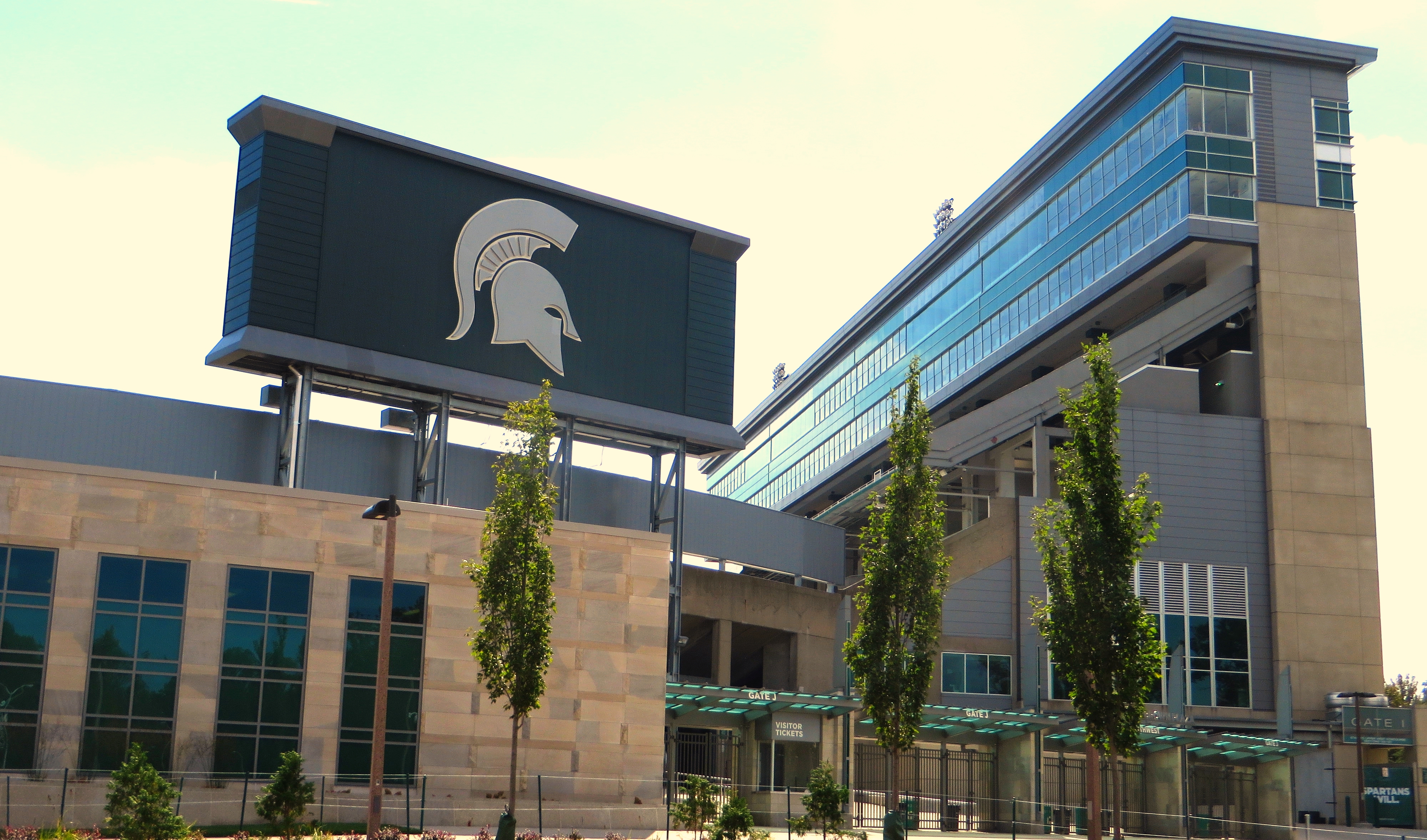 Stadium exterior showing a large video scoreboard with a Spartan helmet logo and a glass and stone facade.
