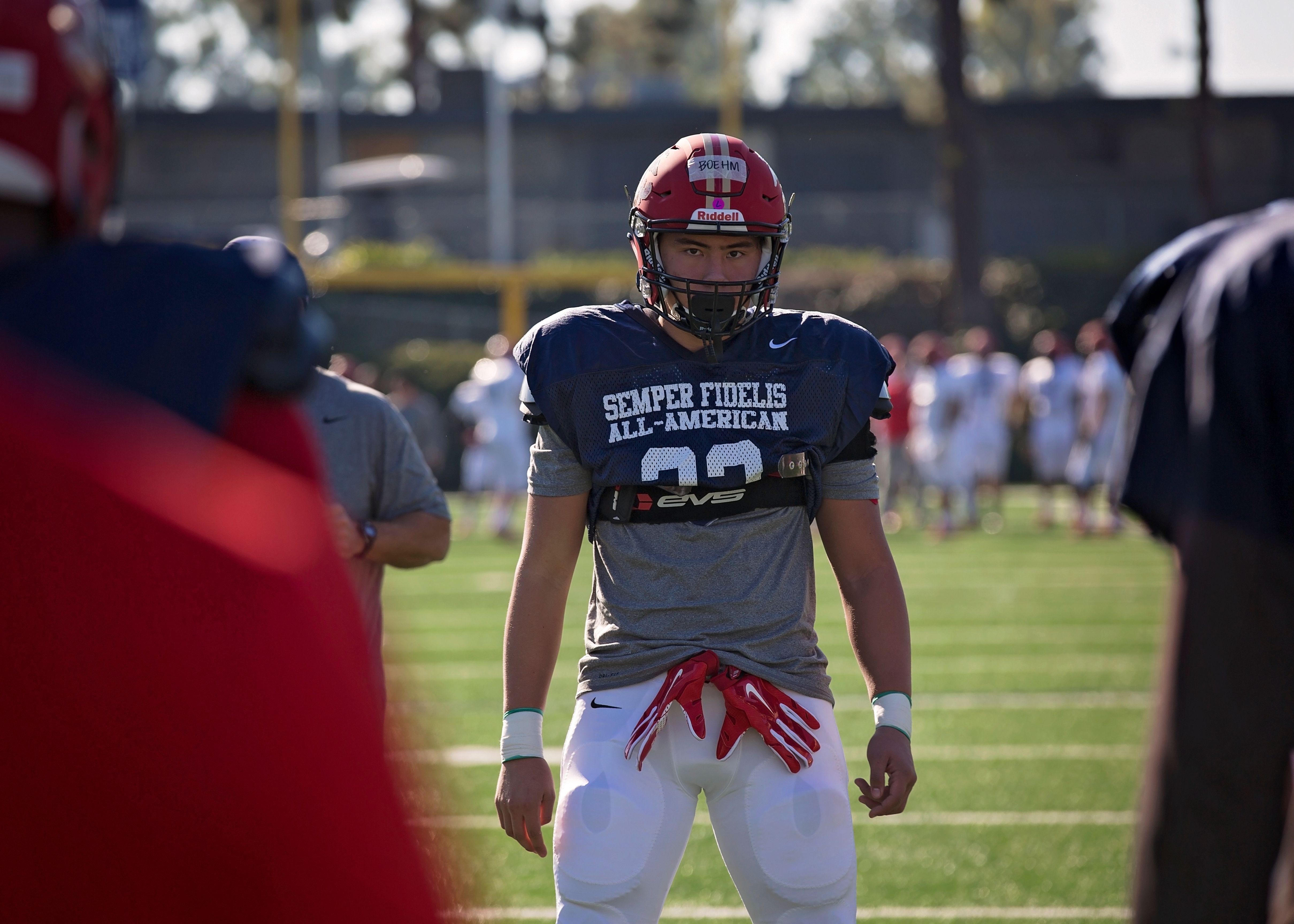 Football player in red helmet and navy jersey stands on a practice field.