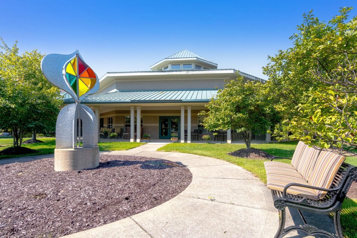 Low-rise building with green metal roof, colorful sculpture, and curved walkway surrounded by trees.