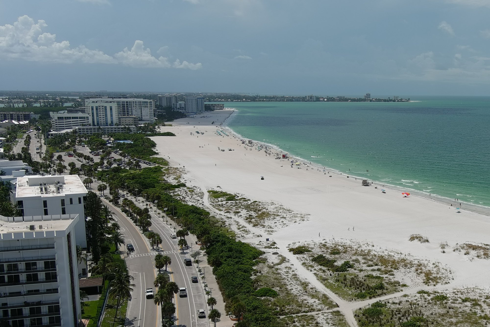Aerial view of a wide white-sand beach alongside turquoise Gulf water and a palm-lined road.