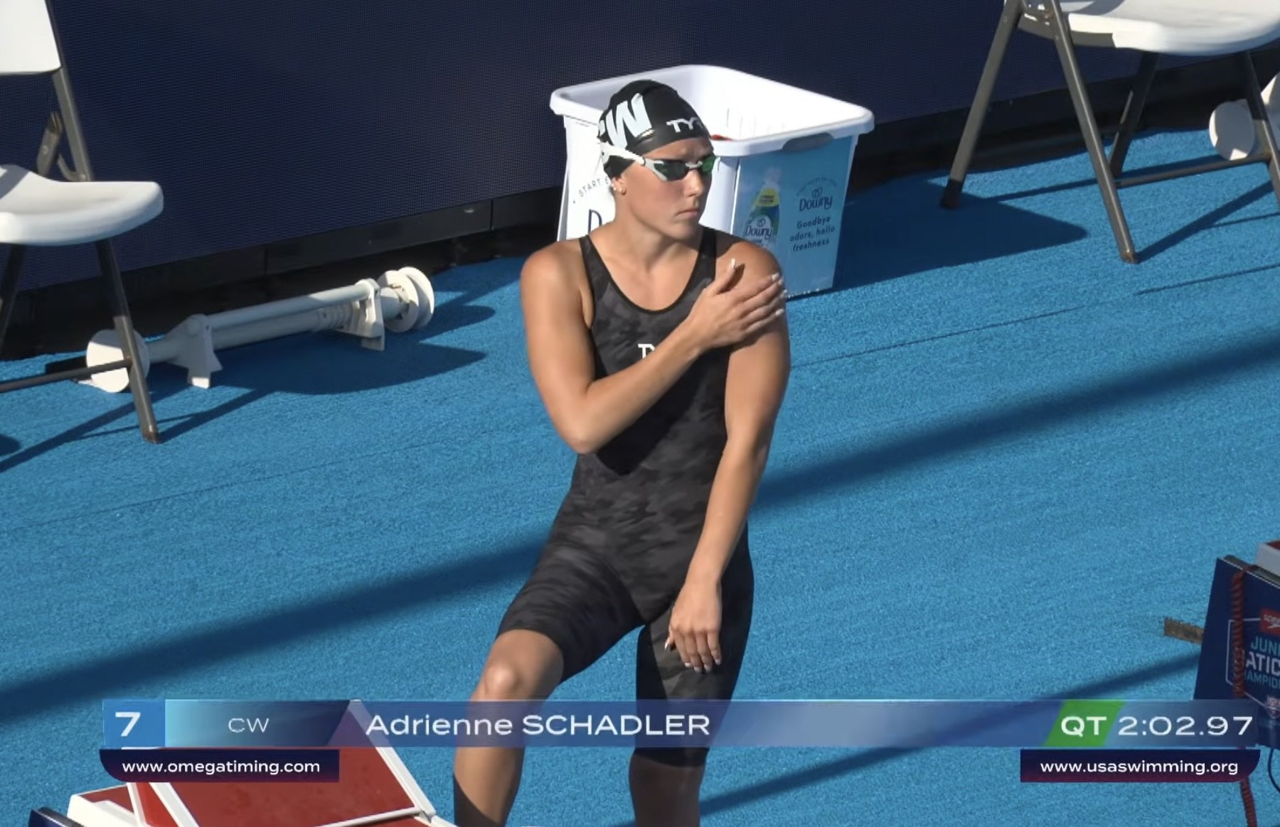 Female swimmer in competition swimsuit and cap stretches poolside with name graphic visible on screen.