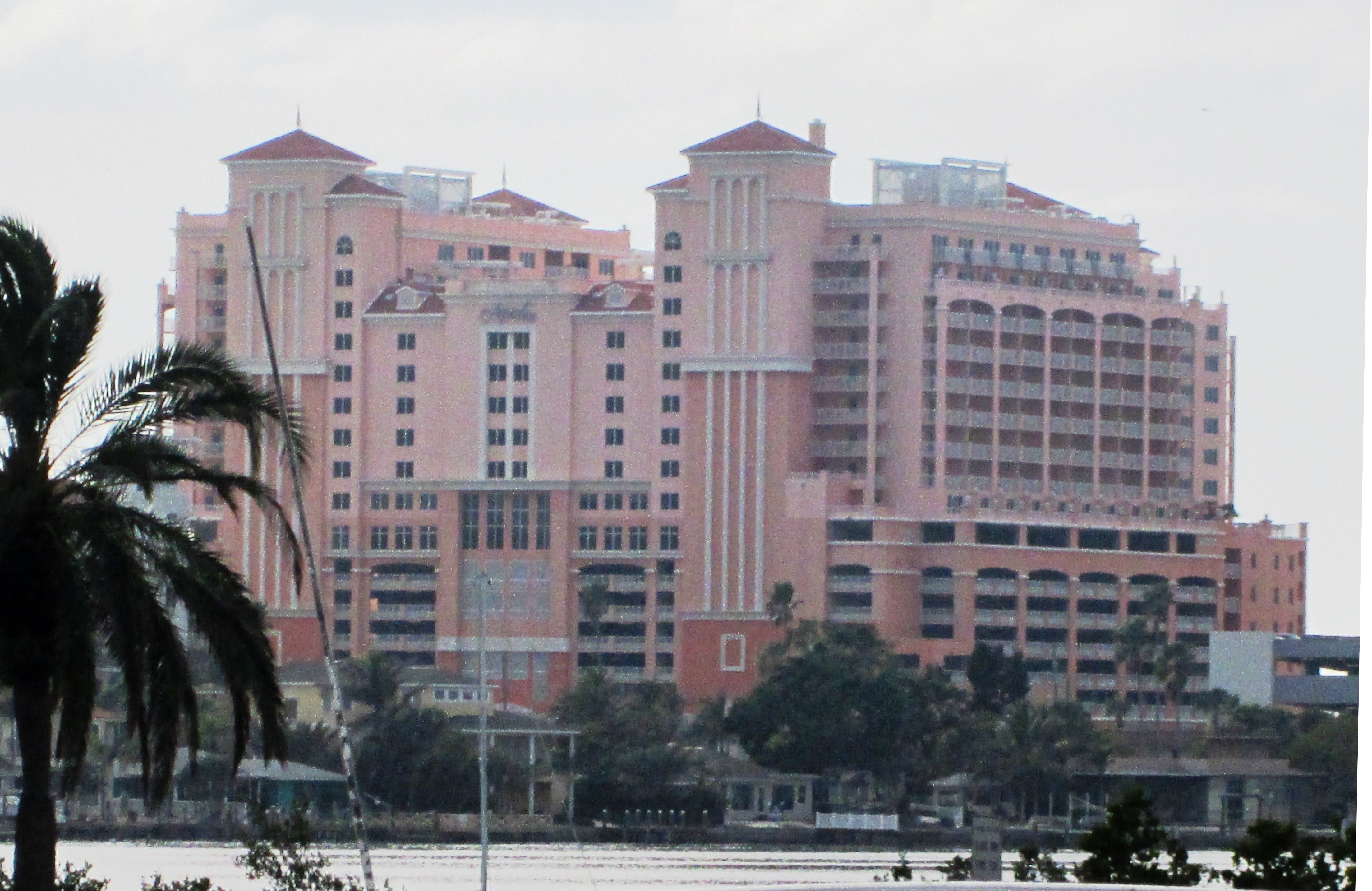 Large pink multi-story resort hotel viewed from across the water with palm trees in the foreground.