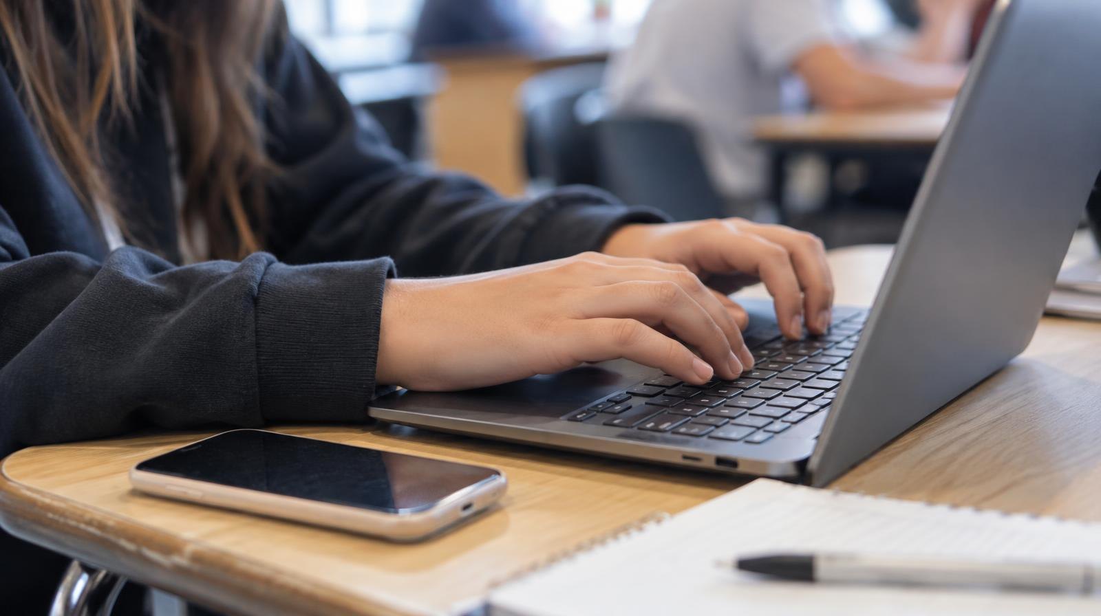 A laptop keyboard and smartphone side by side on a wooden school desk.