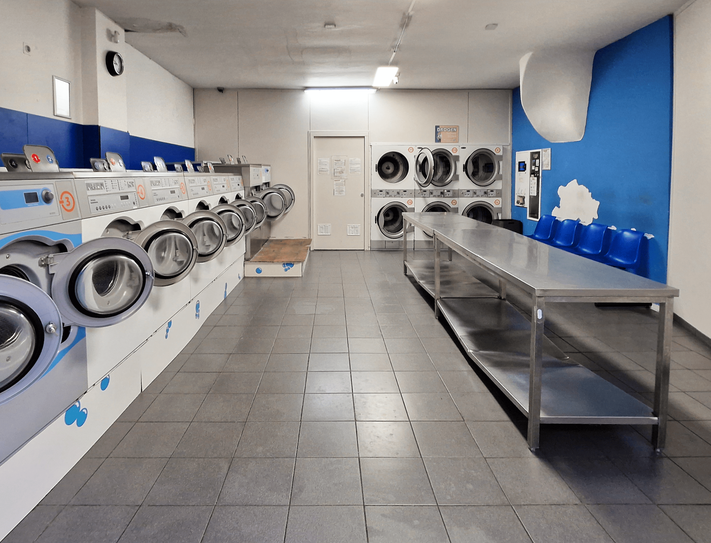 Modern laundromat interior with rows of front-loading washing machines, dryers, and a central folding table.