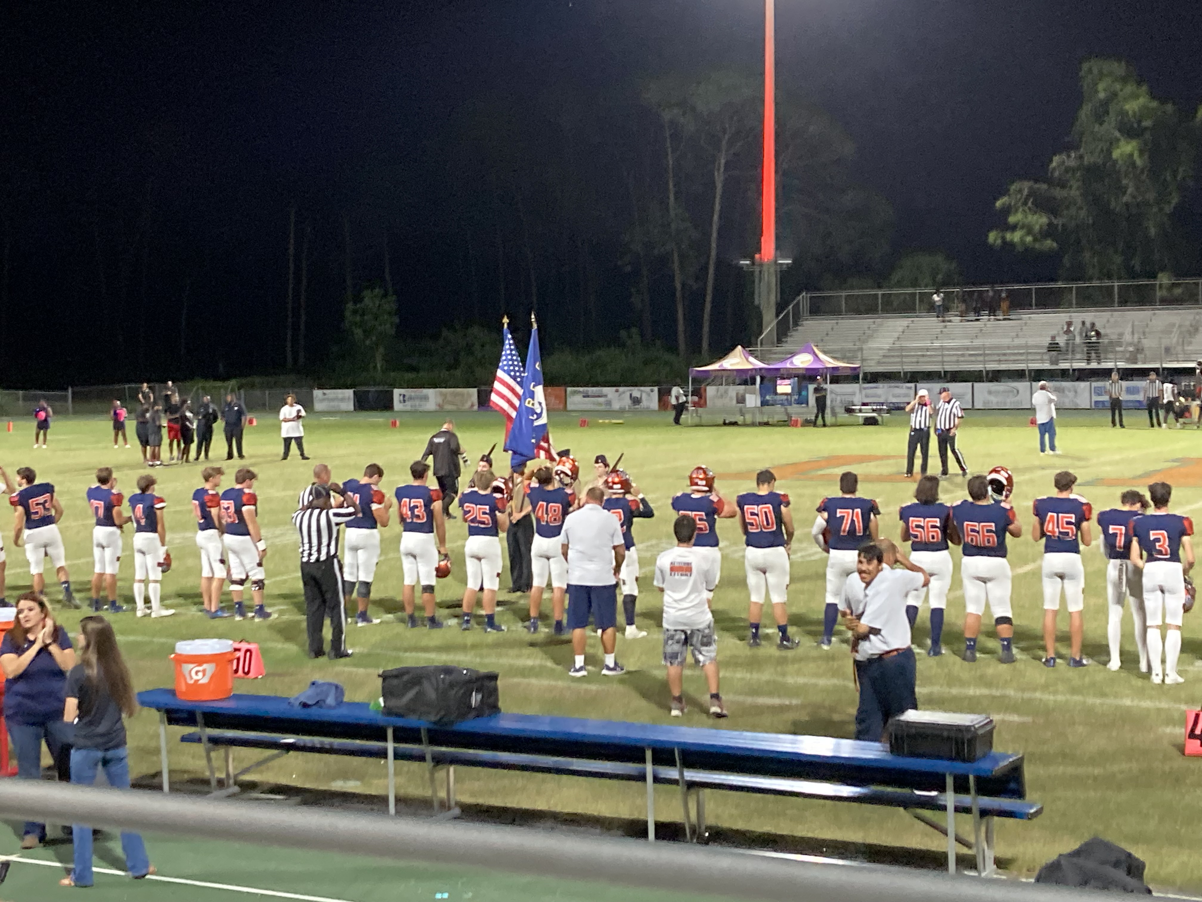 High school football team lined up on a lit field at night during a flag ceremony.