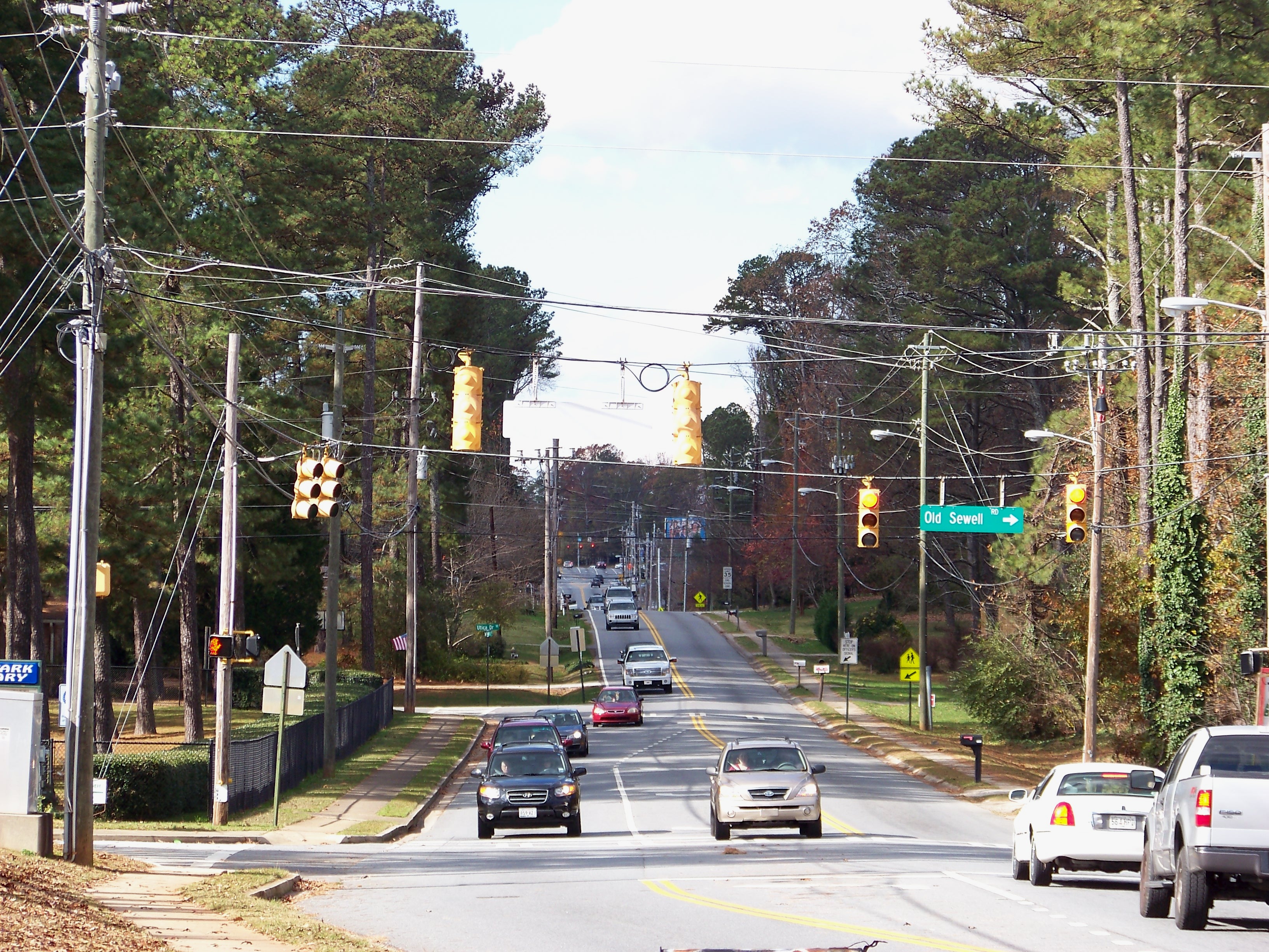 Multi-lane suburban road with traffic signals, utility poles, and trees on both sides.