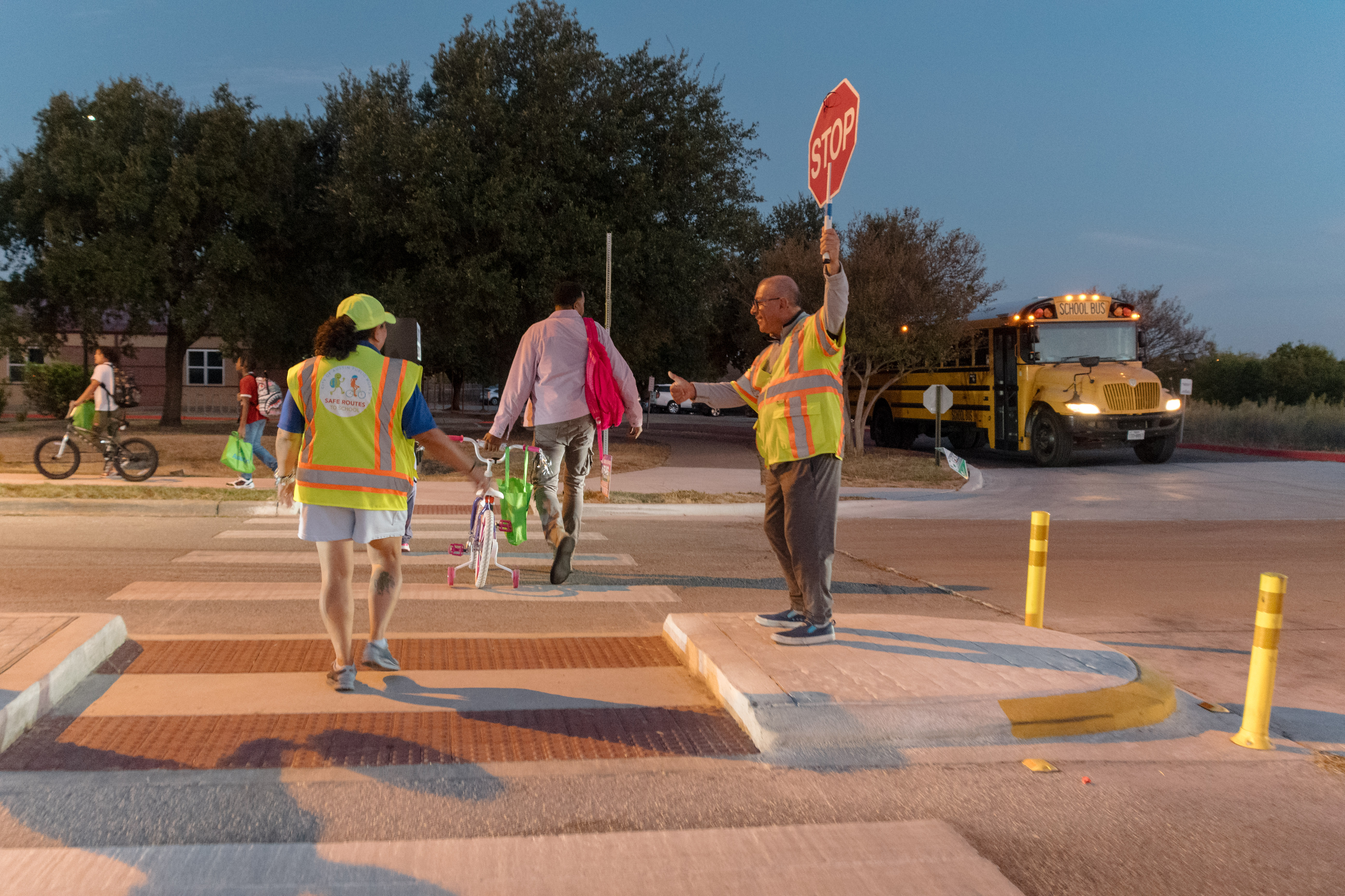 Two crossing guards in yellow vests hold stop signs at a crosswalk with a school bus behind them.