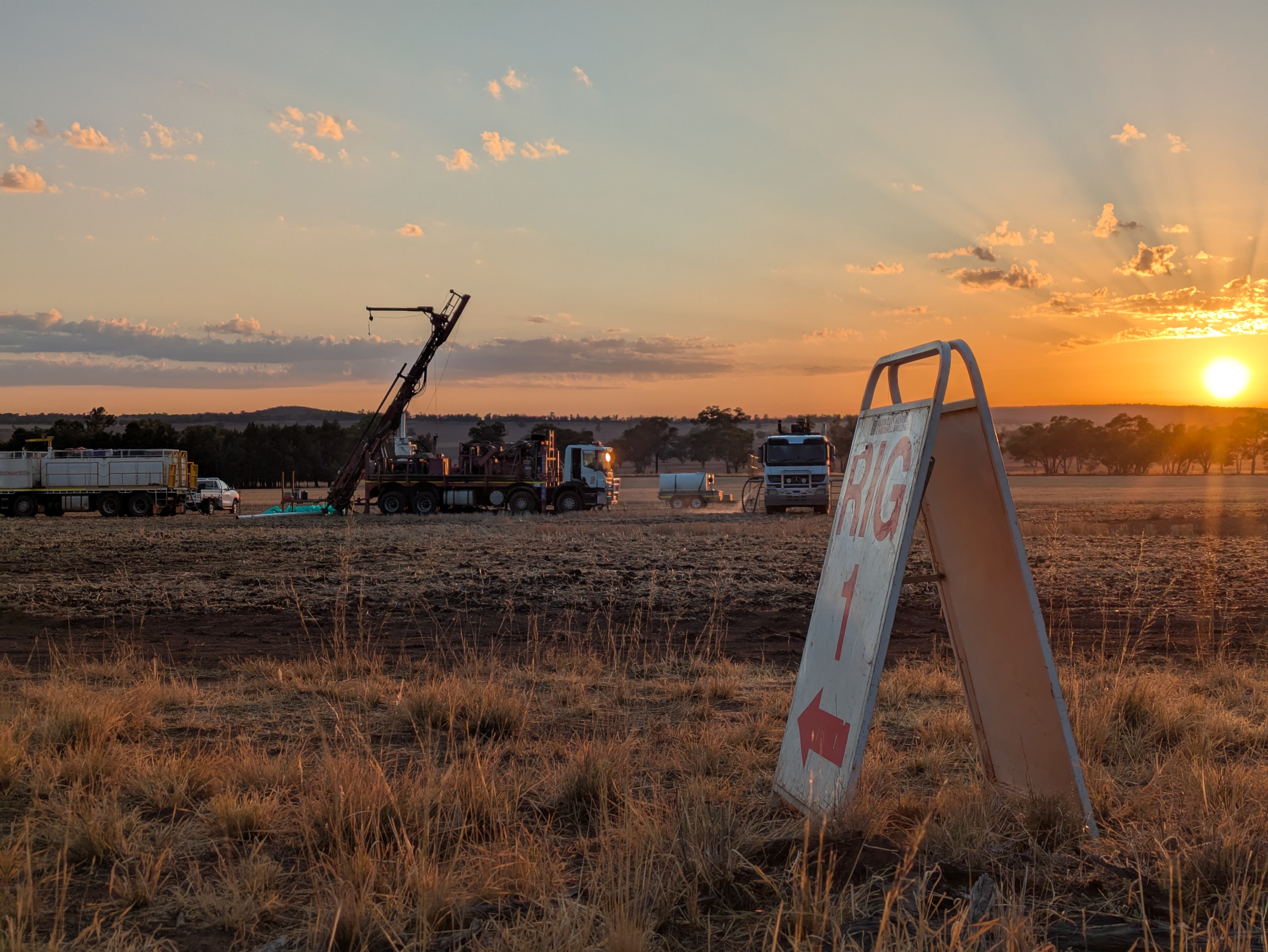 A drilling rig silhouetted against a sunrise sky in an open field.