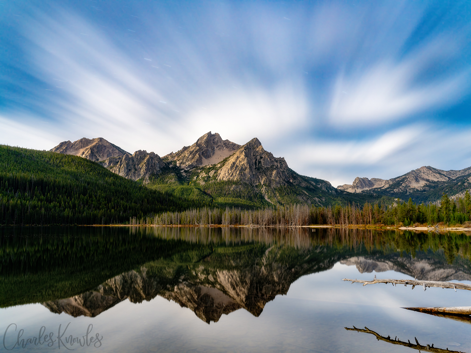 A calm mountain lake reflecting jagged peaks under a moonlit streaked sky with conifer forest along the shore.