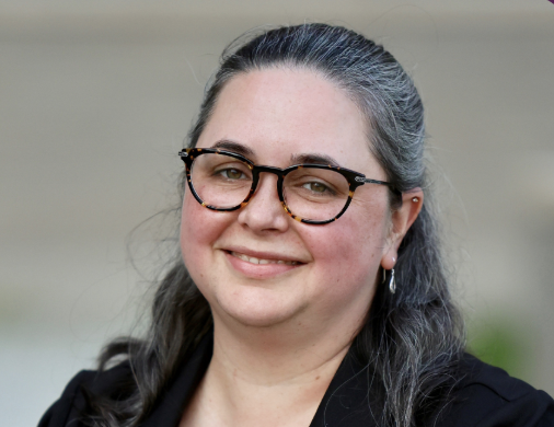Elisabeth Dery smiles, wearing glasses and a black blazer against a blurred background.